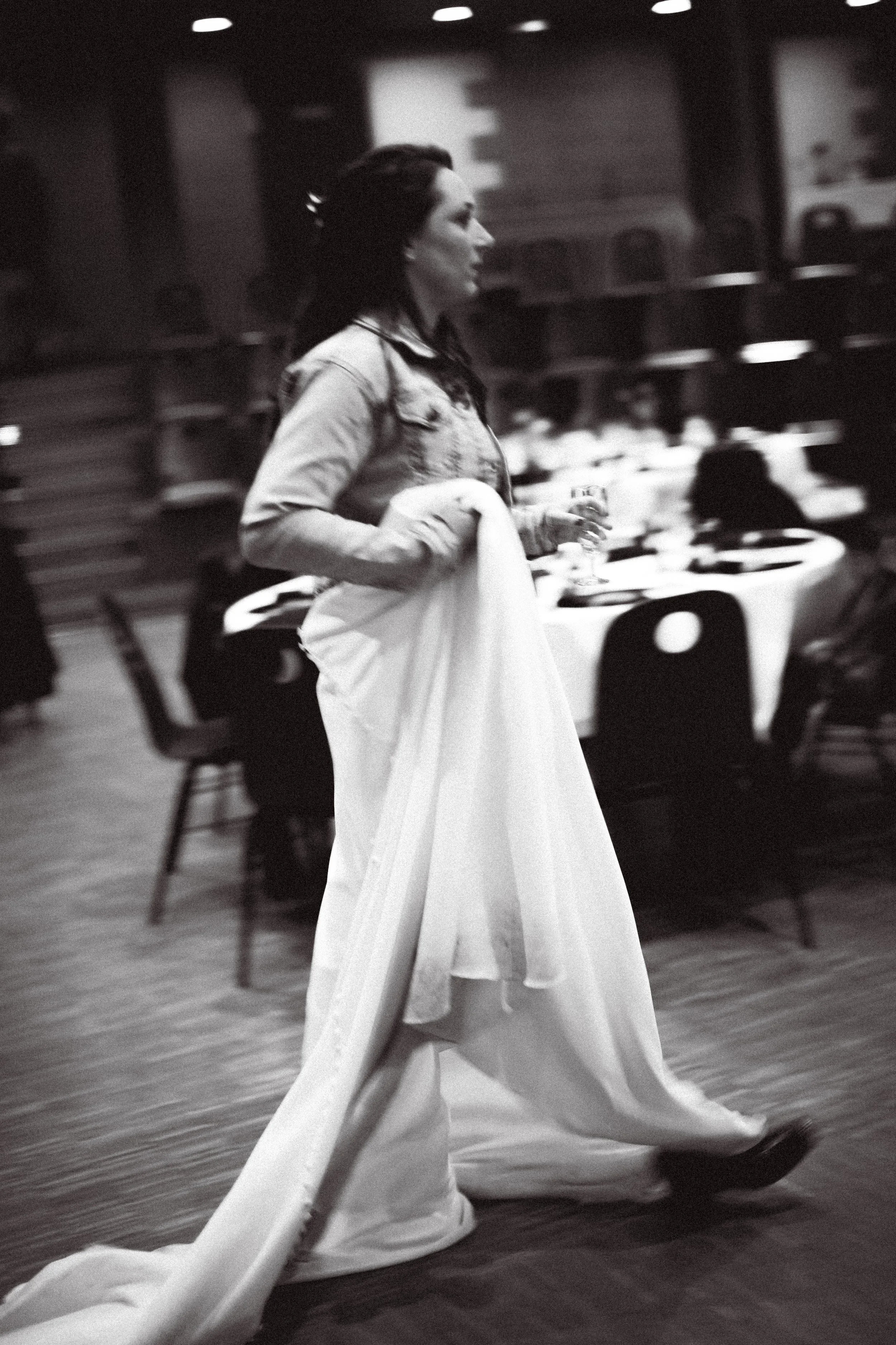 Femme dans une salle de restaurant, portant une robe blanche et une veste, tenant une coupe de vin, en mouvement.