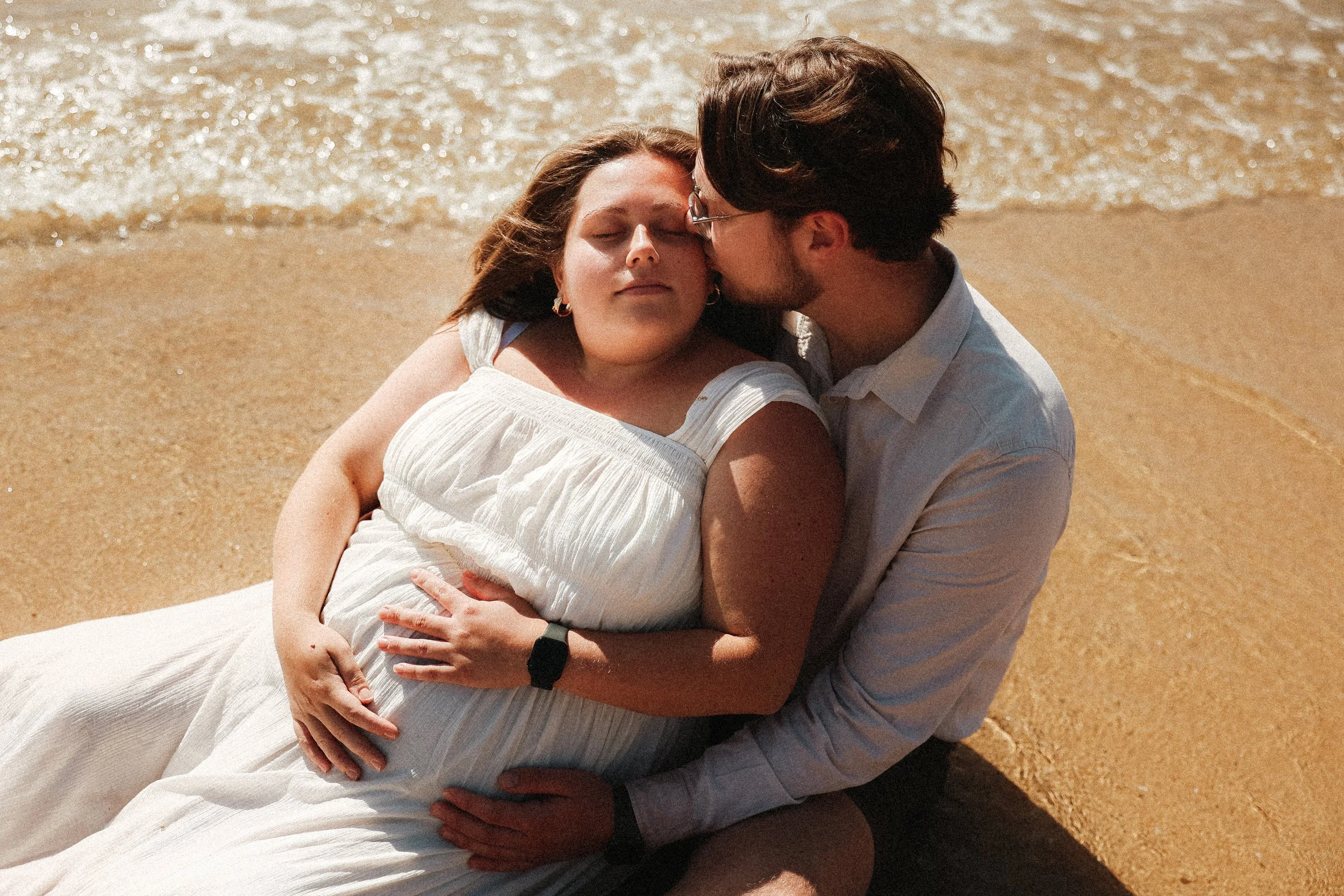 Un couple allongé sur la plage, la femme en robe blanche repose et le homme lui donne un baiser sur le front, près de l'eau.