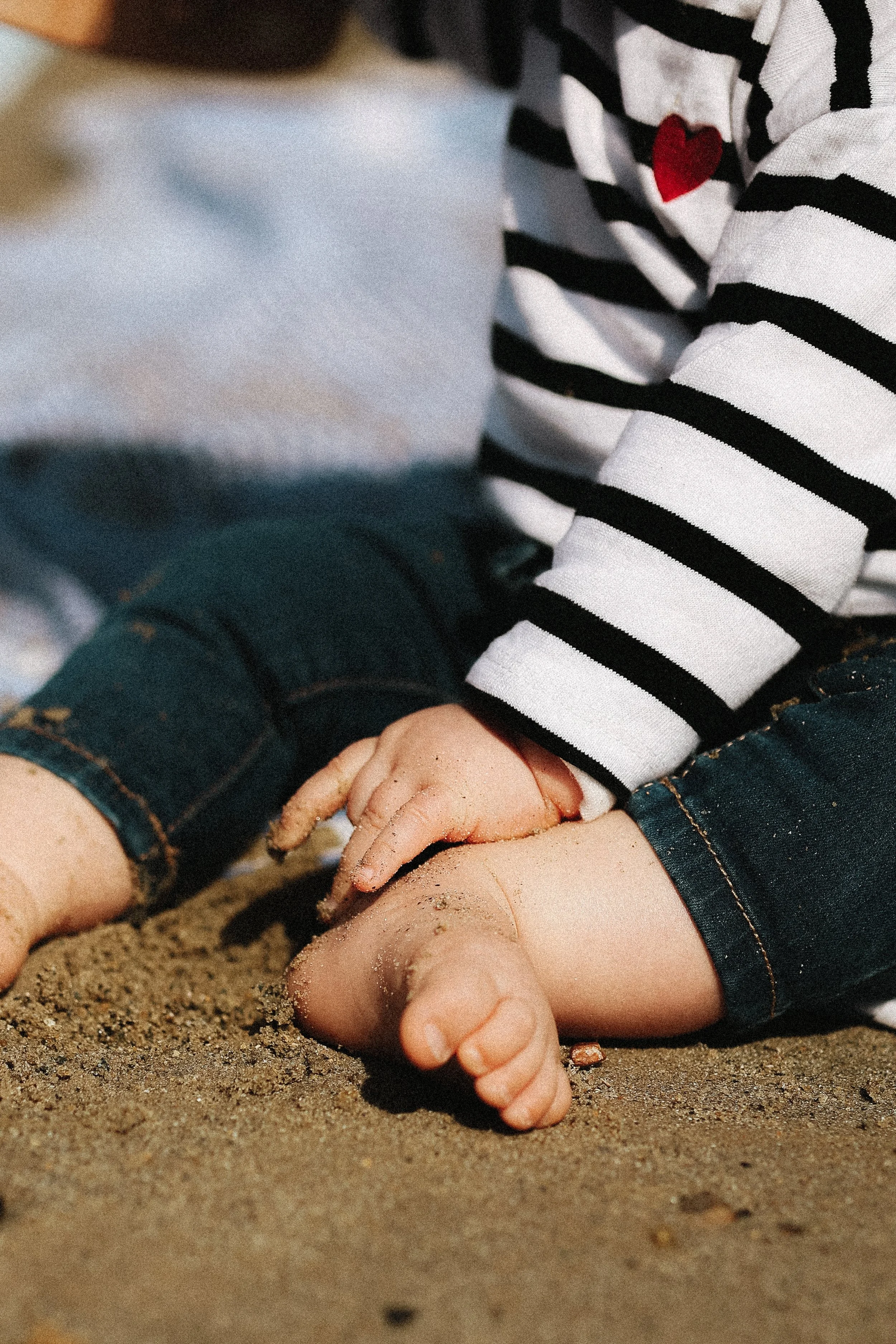 Les mains d'un enfant jouant dans le sable, pieds nus, portant un pantalon en denim et un pull à rayures noires et blanches avec un cœur rouge.