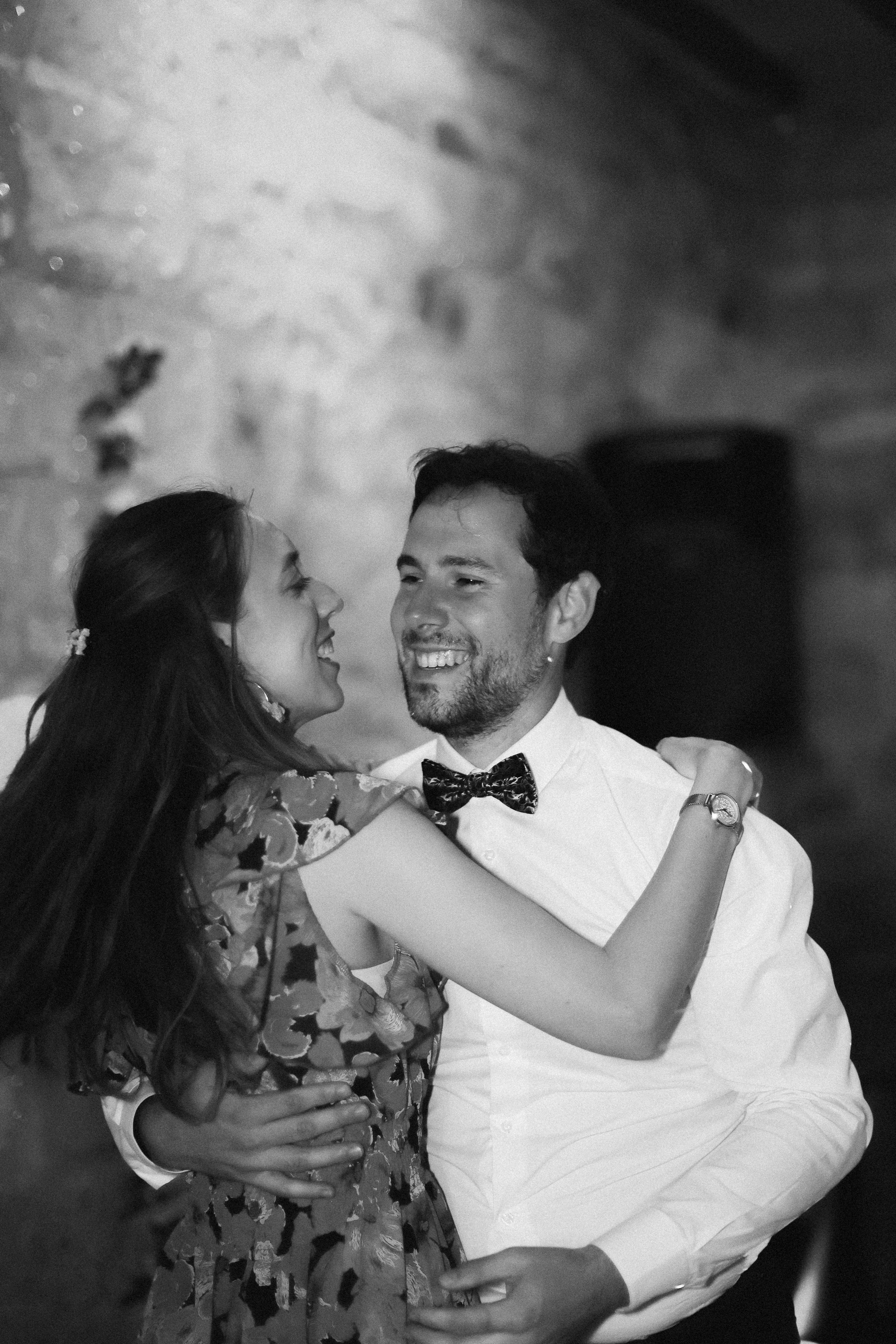 Un couple danse lors d'une occasion spéciale, la femme porte une robe à motifs floraux, et l'homme porte une chemise blanche avec un nœud papillon, souriant.