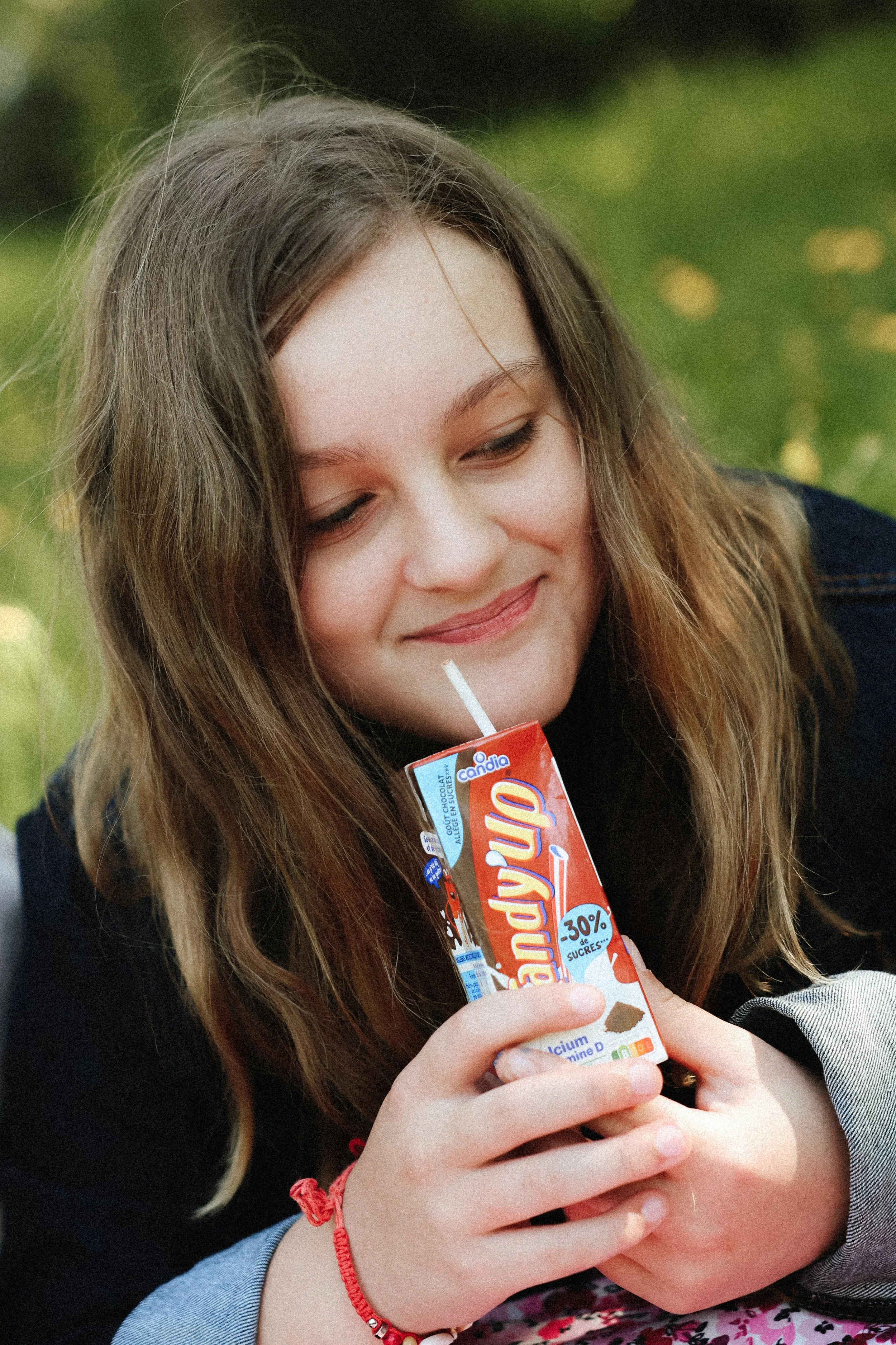 Jeune fille aux cheveux bruns, tenant un paquet de bonbons à la nicotine sans sucre, souriant légèrement, dans un environnement extérieur verdoyant.