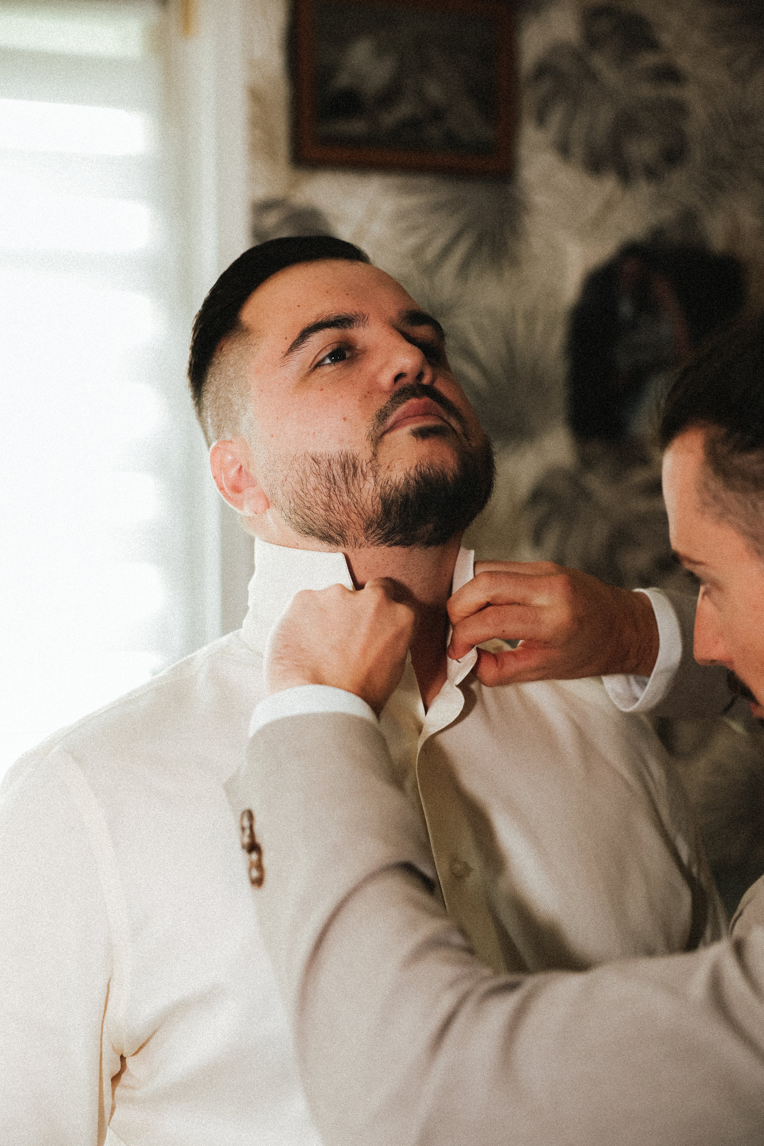 Un homme en chemise blanche est aidé à boutonner son col par une autre personne. Il a une barbe et des cheveux foncés, dans un intérieur avec une fenêtre lumineuse et un décor mural.