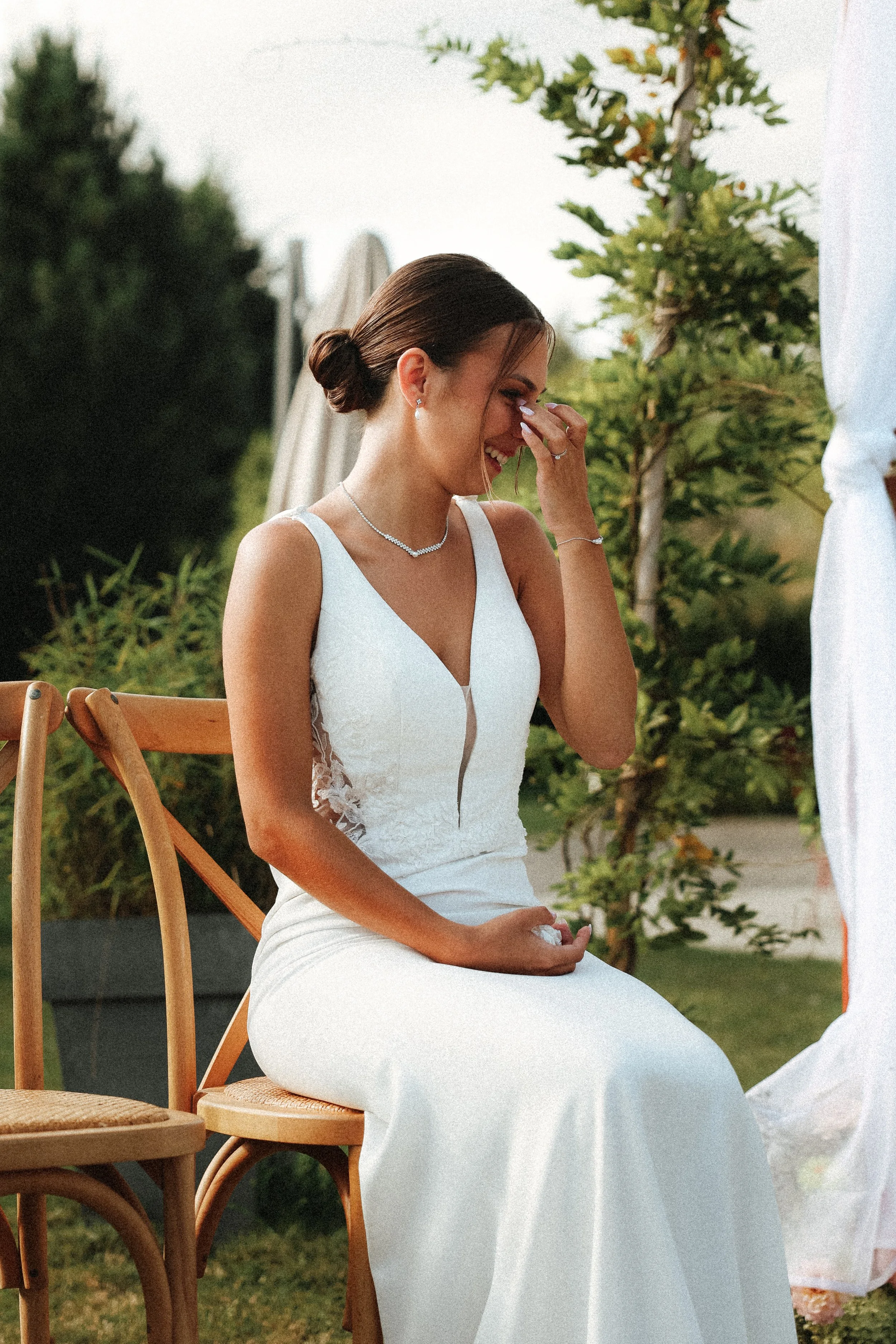 Une jeune femme en robe blanche est assise sur une chaise en bois lors d'une cérémonie, souriant tout en se tenant la main et en levant la main vers son visage, dans un environnement extérieur avec des arbustes et un ciel clair.