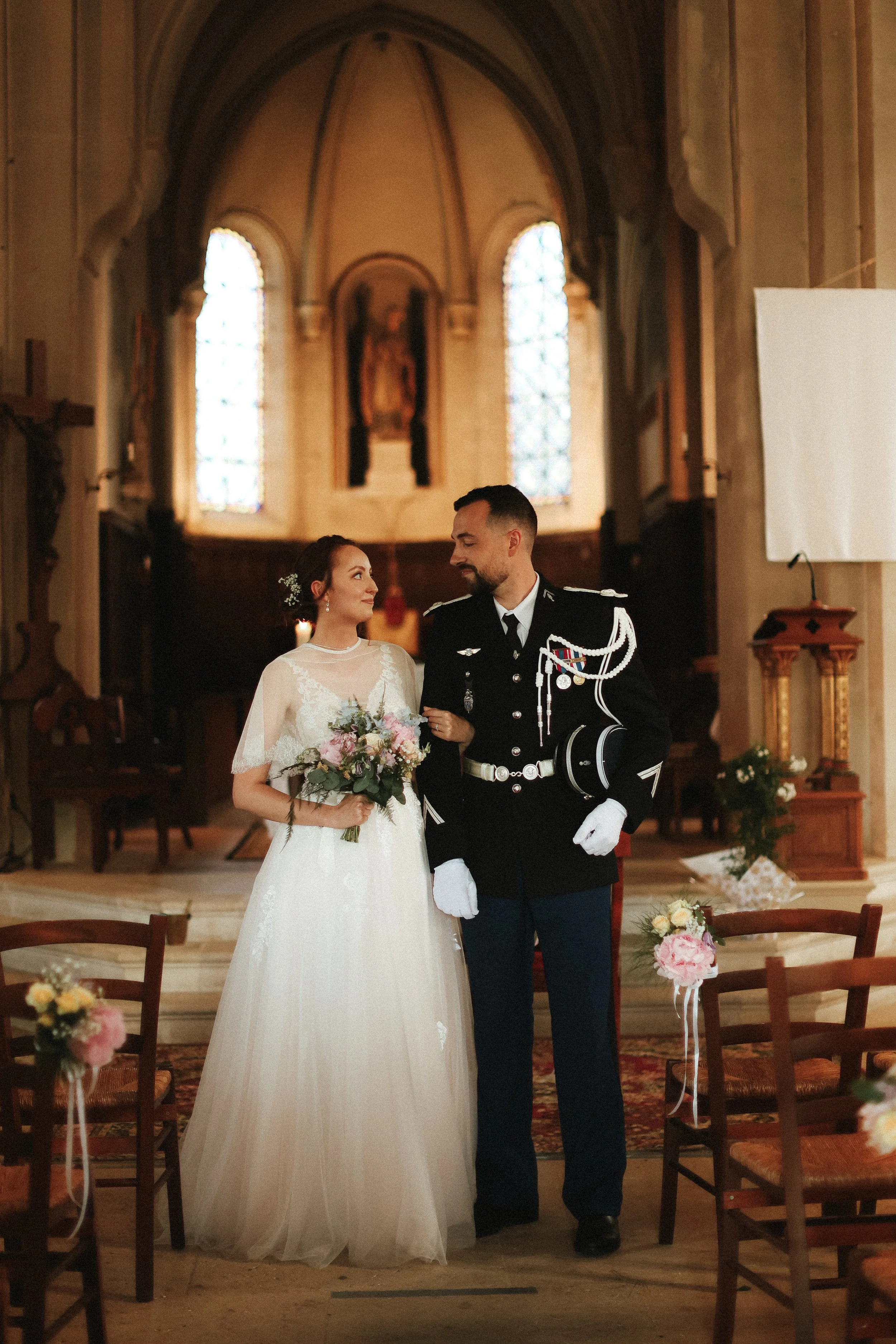 Un couple de mariés dans une église, la femme en robe blanche tenant un bouquet de fleurs, l'homme en uniforme militaire, se regardant tendrement.