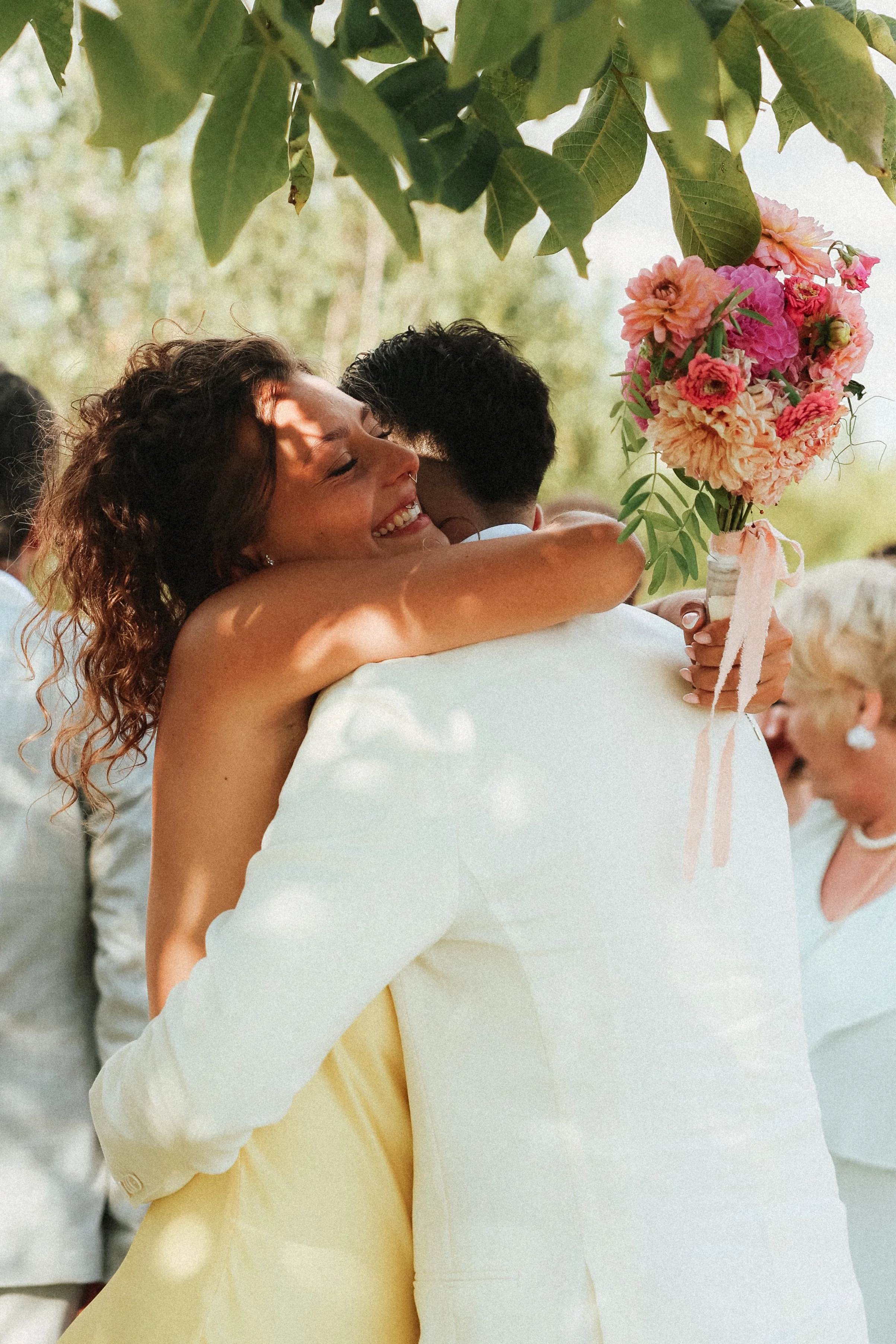 Deux personnes se font un câlin lors d'une cérémonie en plein air. La femme sourit et tient un bouquet de fleurs colorées. Il y a d'autres personnes en arrière-plan.