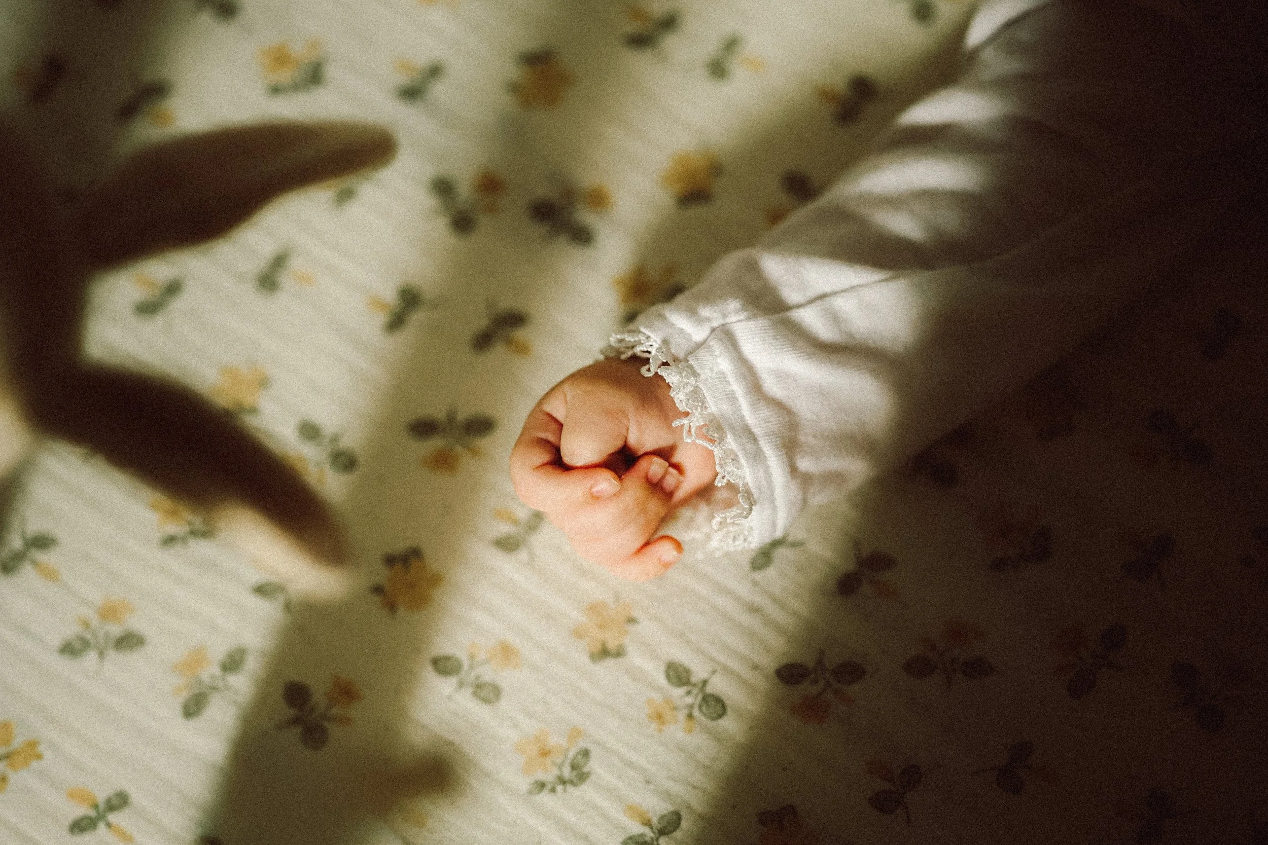 Une main de bébé avec une manche en dentelle blanche, faisant un geste de poing, avec une lumière douce et une ombre sur un tissu à motifs floraux.