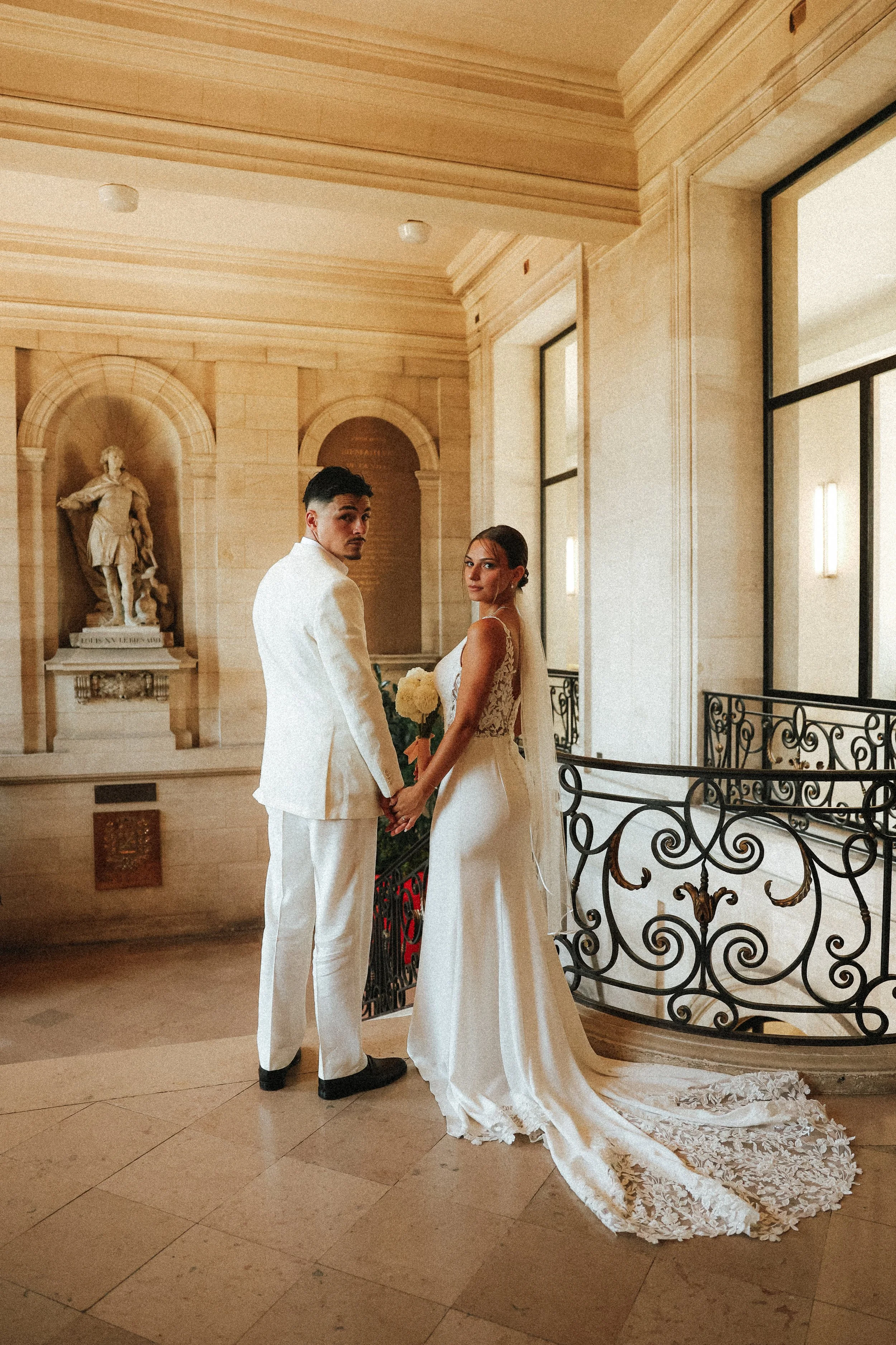 Un couple de mariés en robes blanches se tenant la main dans une salle élégante avec des murs en pierre et des fenêtres grandes. La mariée porte une robe de mariage avec un traîneau en dentelle, le marié porte un costume blanc.