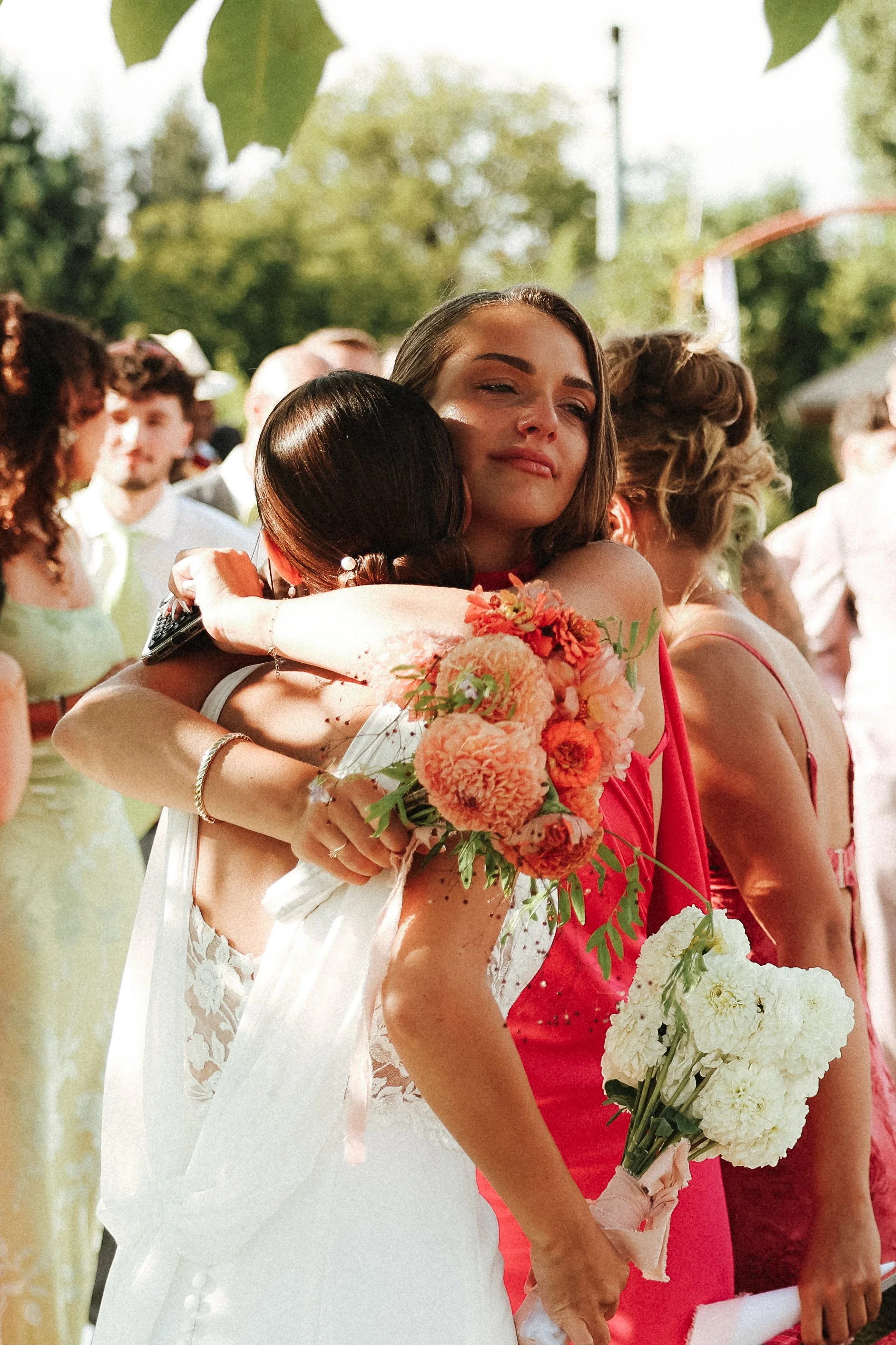 Deux femmes s'embrassent lors d'une cérémonie en plein air, entourées d'autres personnes, avec une végétation en arrière-plan.