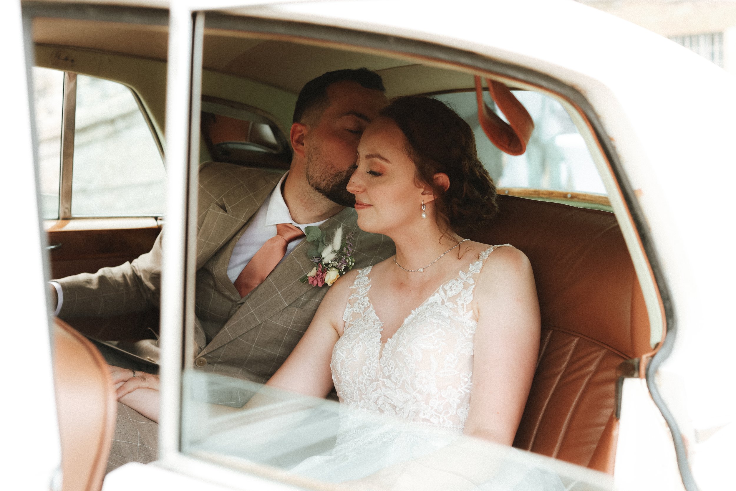 Un couple de mariés dans une voiture ancienne, lui embrasse la joue de la mariée.