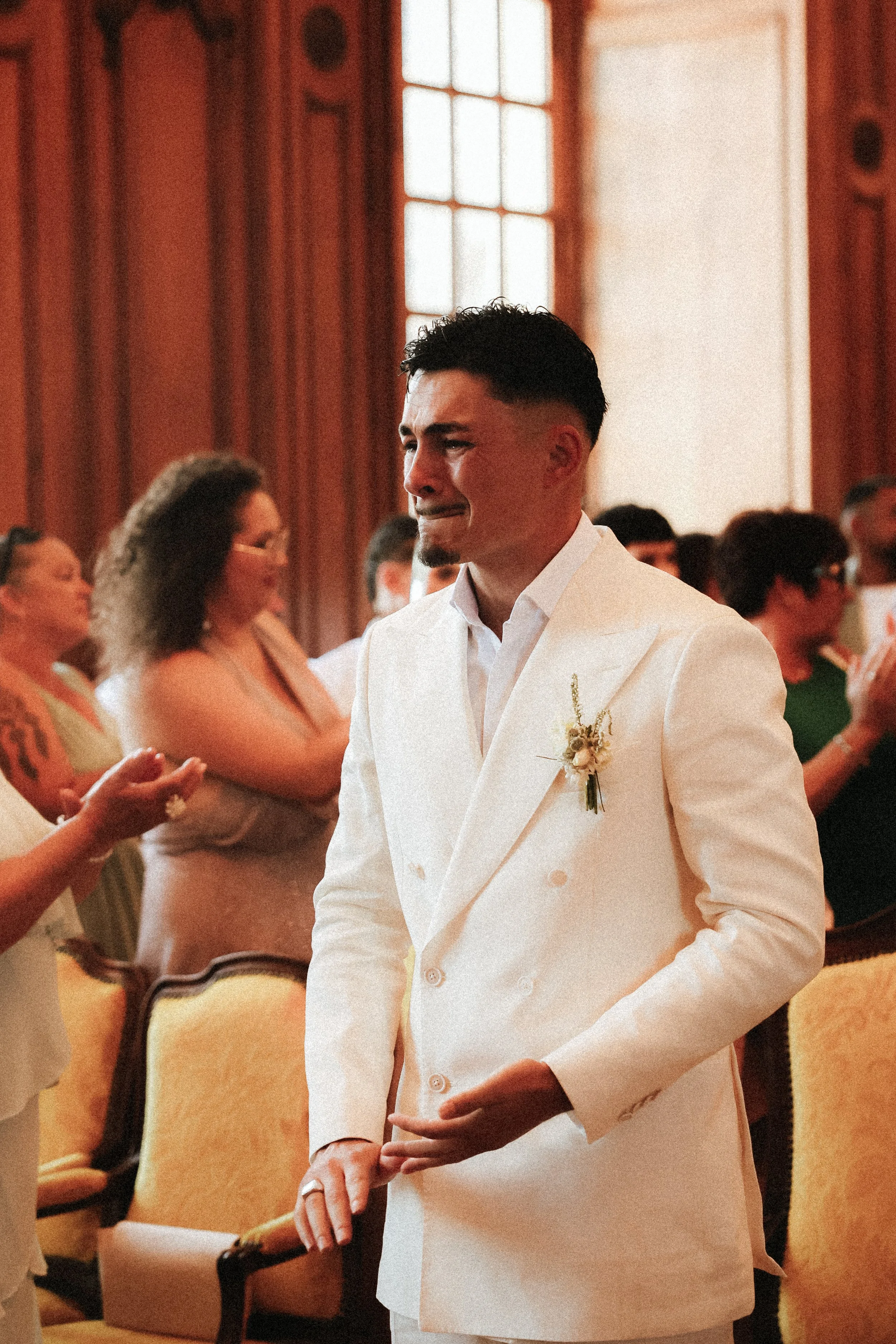 Un homme en costume blanc lors d'une cérémonie, entouré de plusieurs personnes dans un endroit élégant avec des fenêtres en bois et des fauteuils orangés.