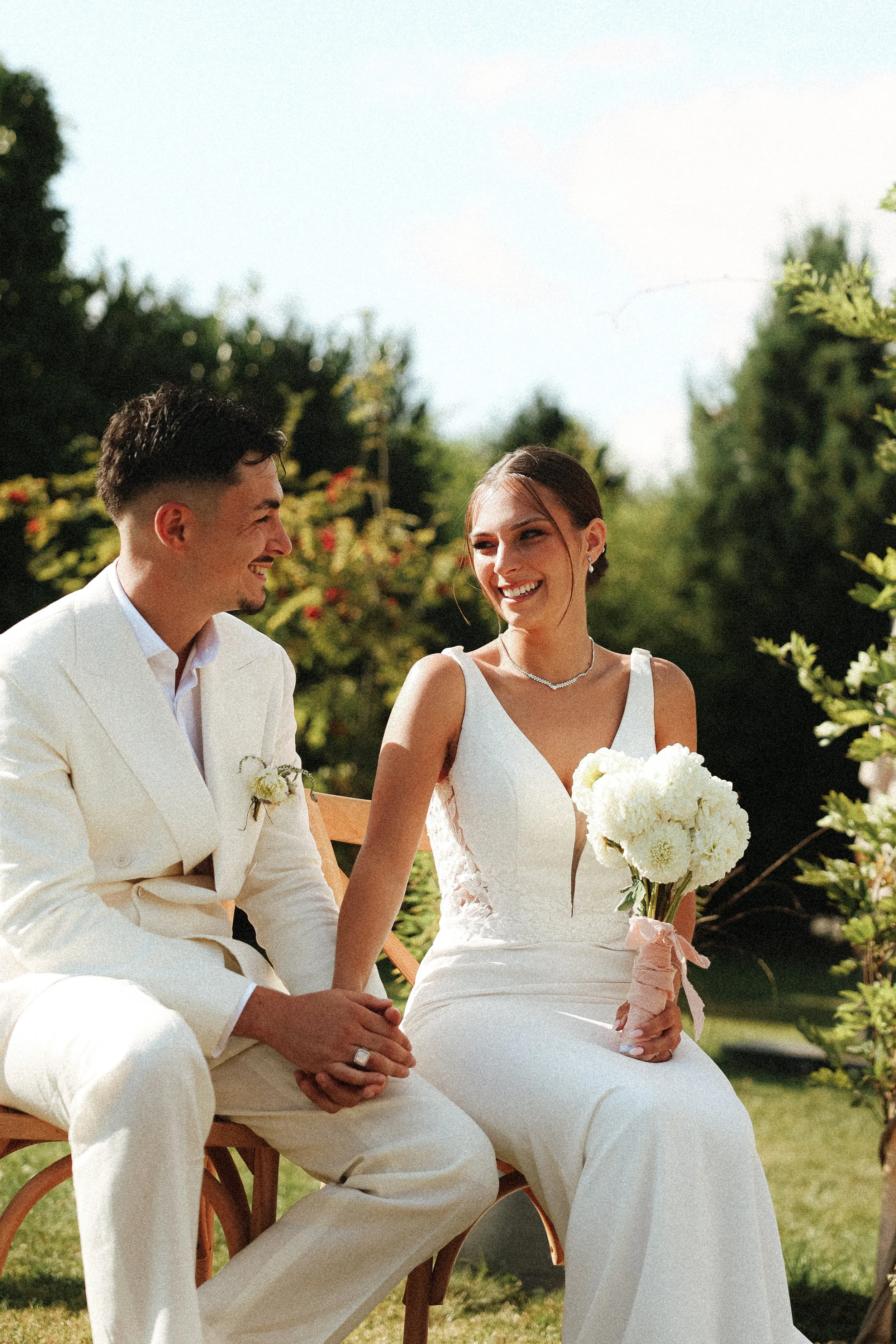 Un couple en robes de mariage assis à l'extérieur, la femme tenant un bouquet de fleurs blanches, souriant lors d'une cérémonie de mariage en plein air.