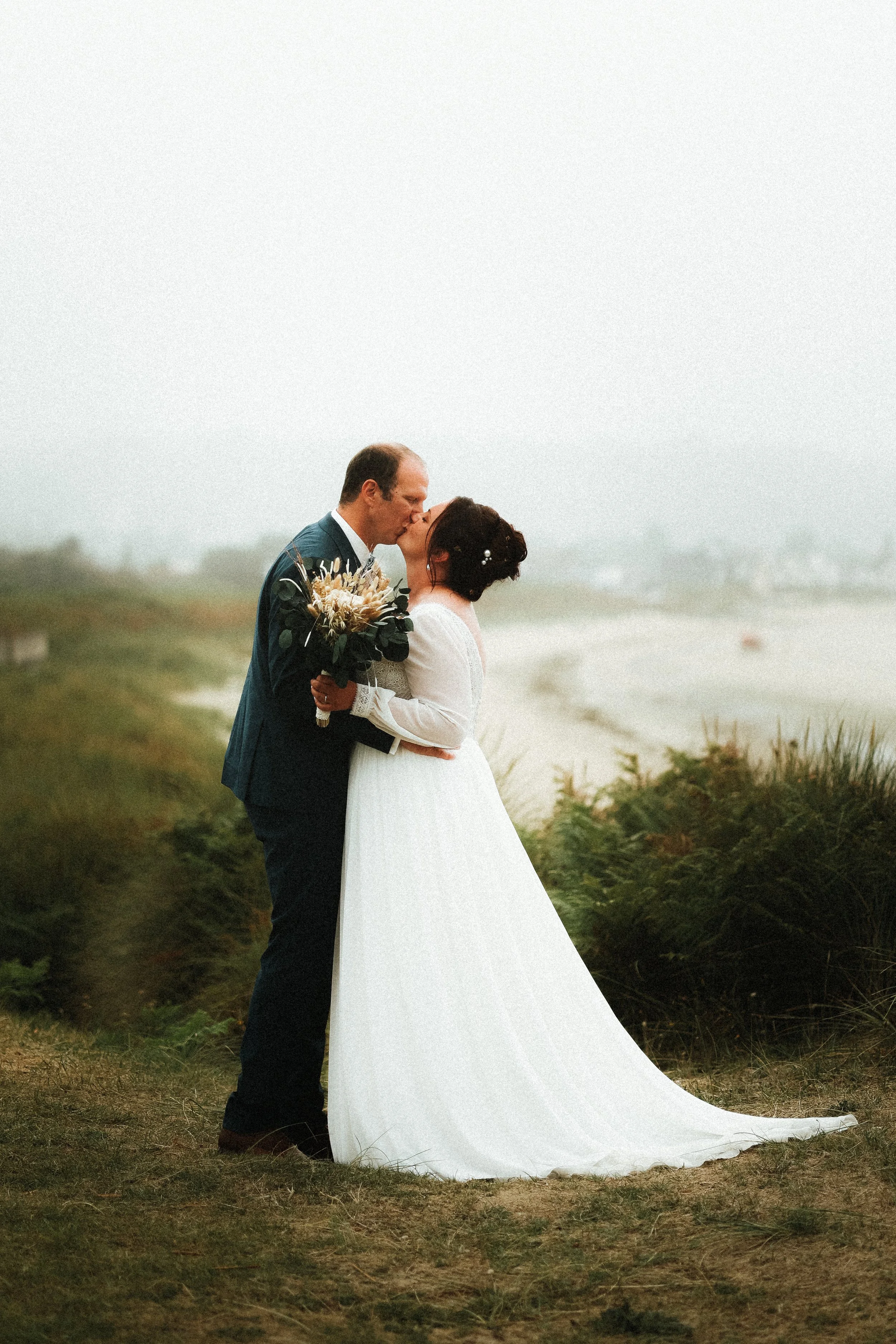 Un couple de mariés s'embrassant lors d'une séance photo en extérieur, devant la mer et la côte.