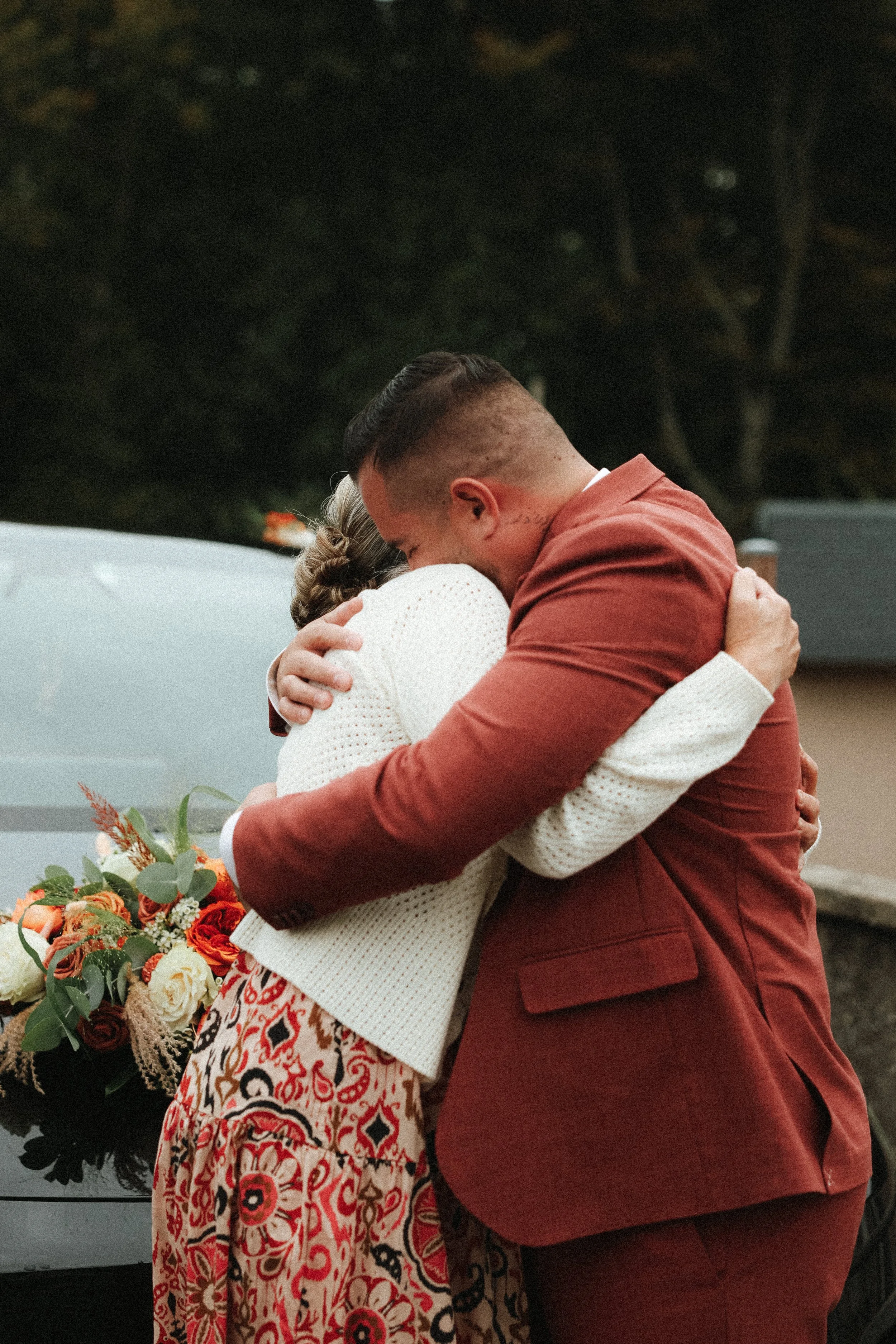 Deux personnes s'embrassent en un câlin dans un cadre extérieur, avec un bouquet de fleurs à côté.
