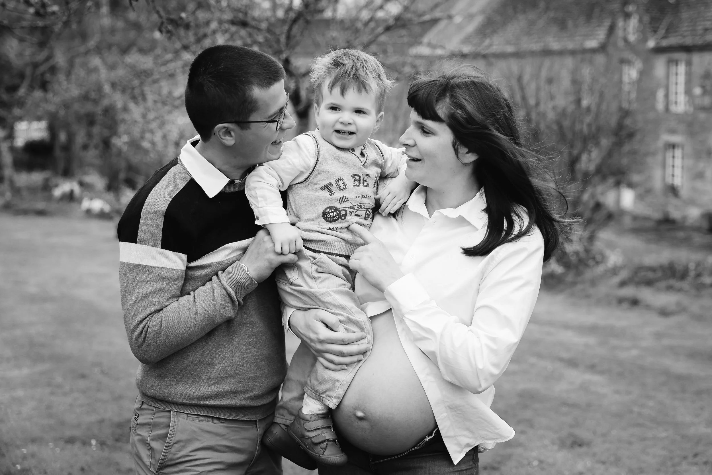 Une famille souriante composée d'un homme, d'une femme enceinte et d'un petit garçon, tous dehors dans un parc.