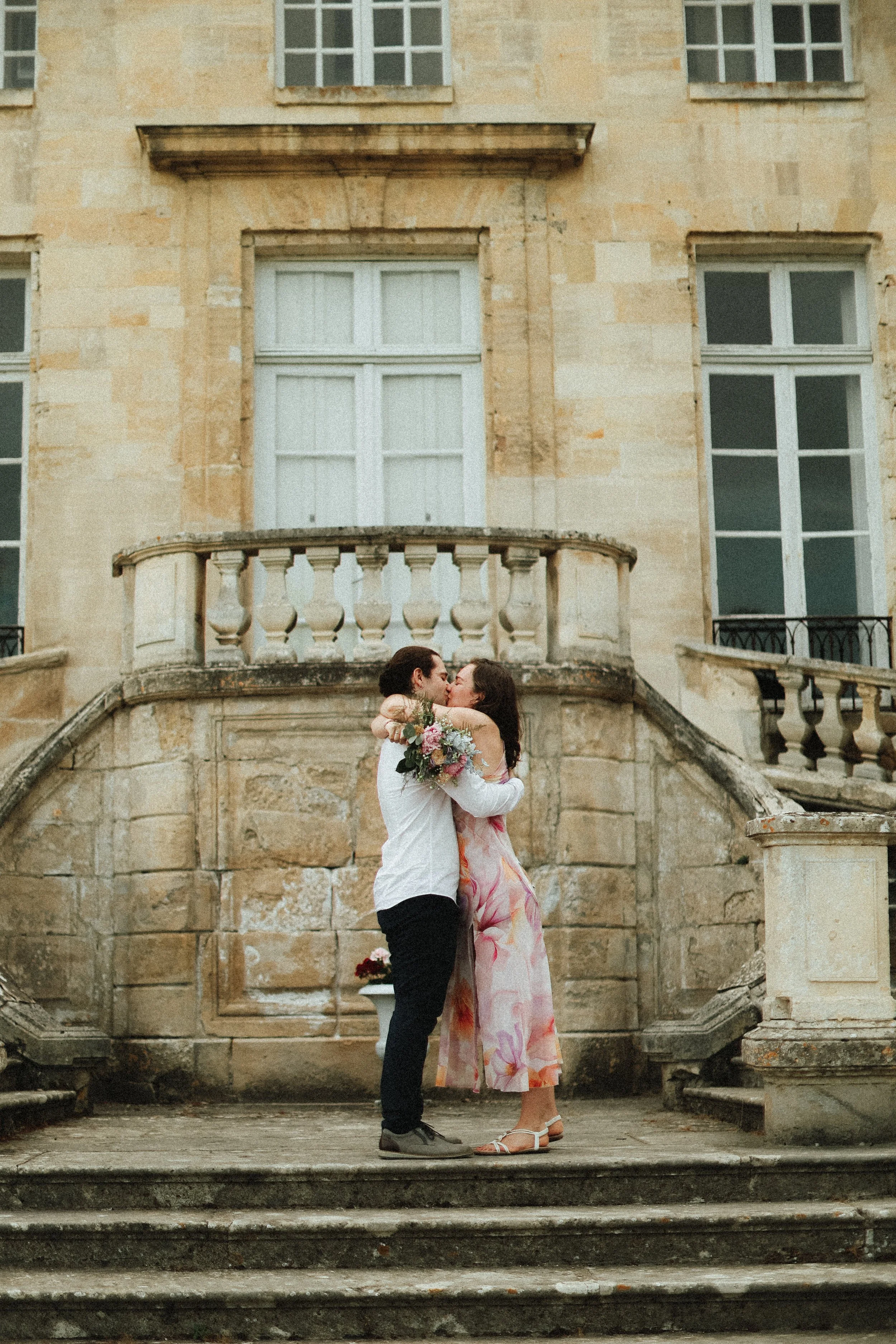 Un couple s'embrasse en haut des escaliers devant un bâtiment ancien en pierre, avec la femme tenant un bouquet de fleurs.