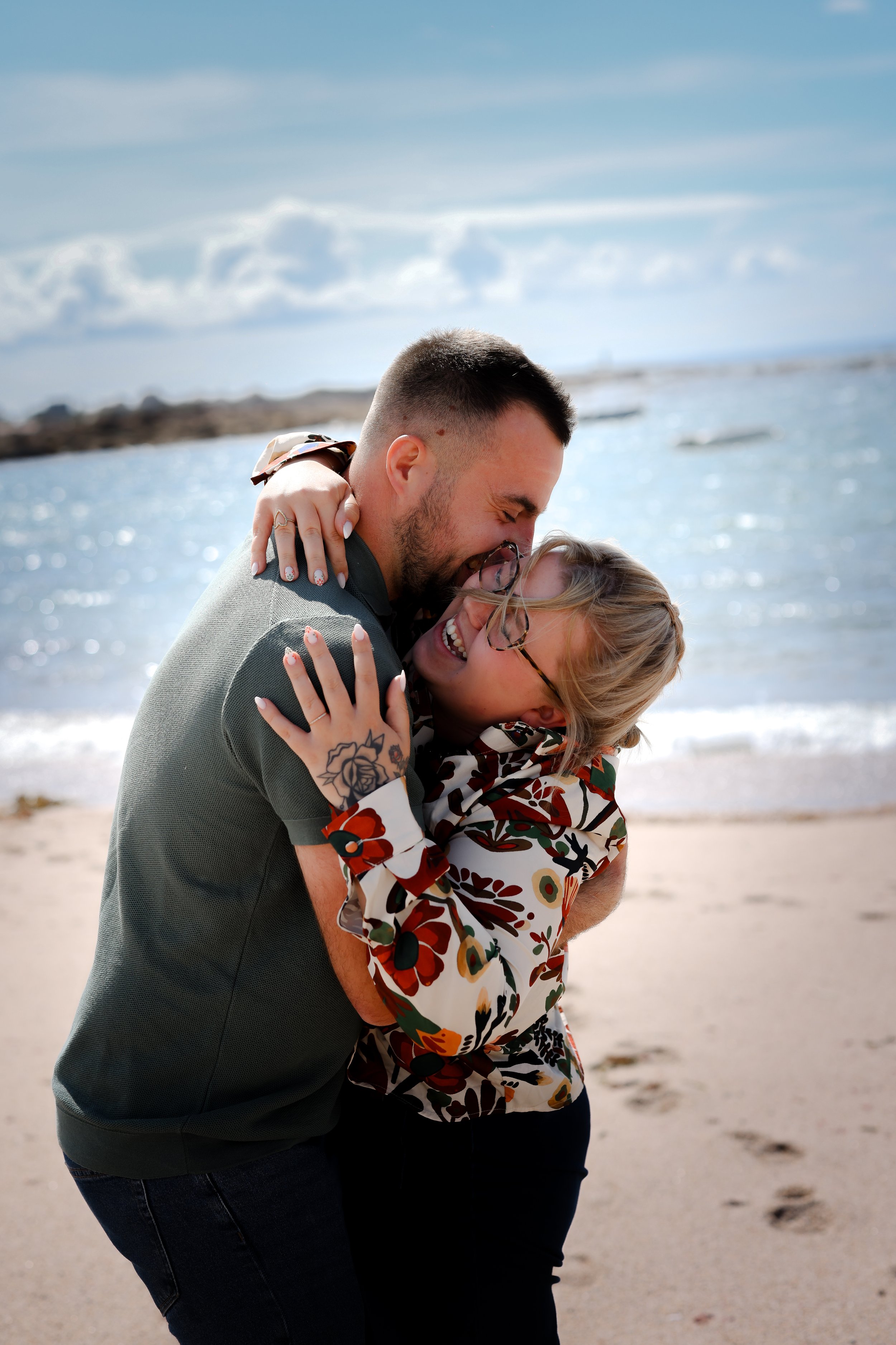 Un couple s'embrasse et rit sur la plage, avec la mer et un ciel bleu en arrière-plan.
