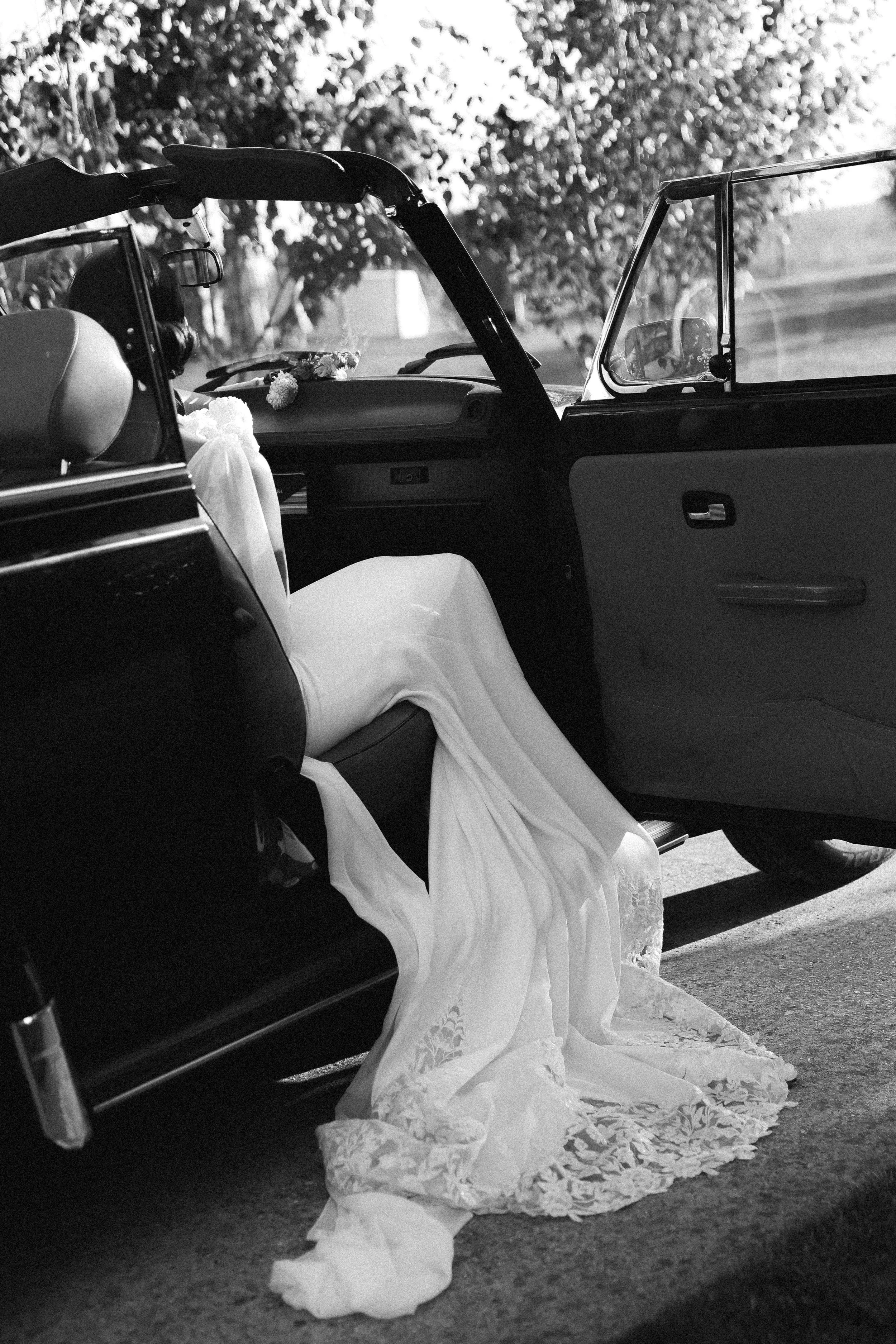 Une voiture avec une robe de mariée suspendue à l'intérieur, vue de l'intérieur avec le porte-voiture ouvert, en noir et blanc.