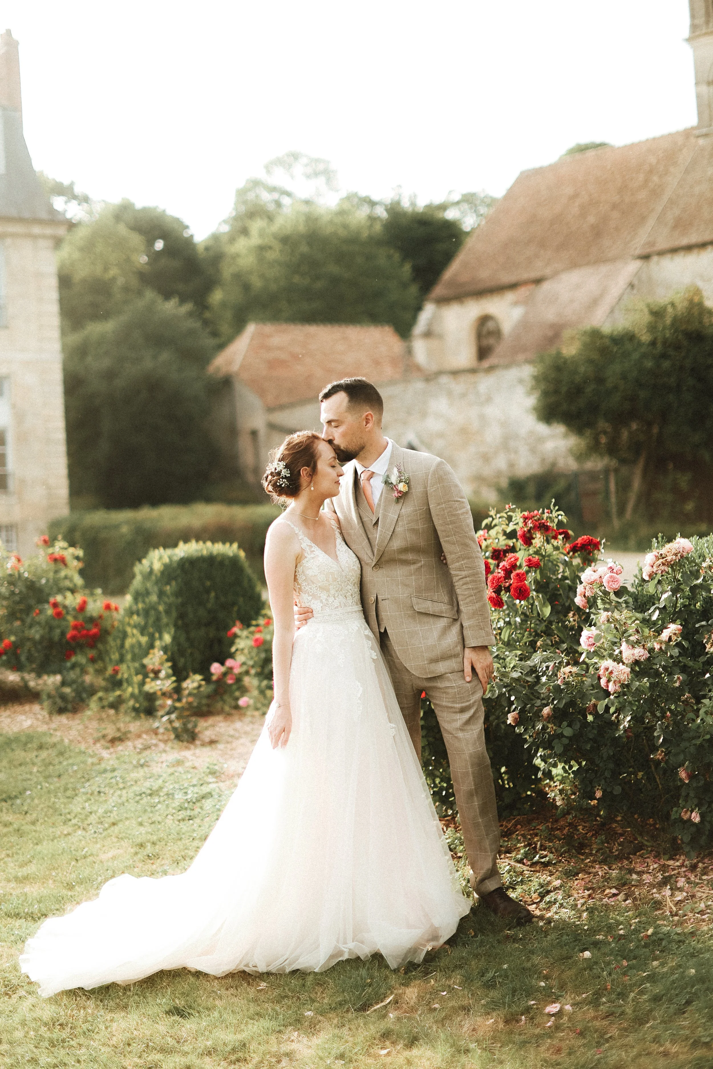 Un couple de mariés posant dans un jardin fleuri, avec une vieille église en pierre en arrière-plan, le mari embrassant tendrement la mariée.