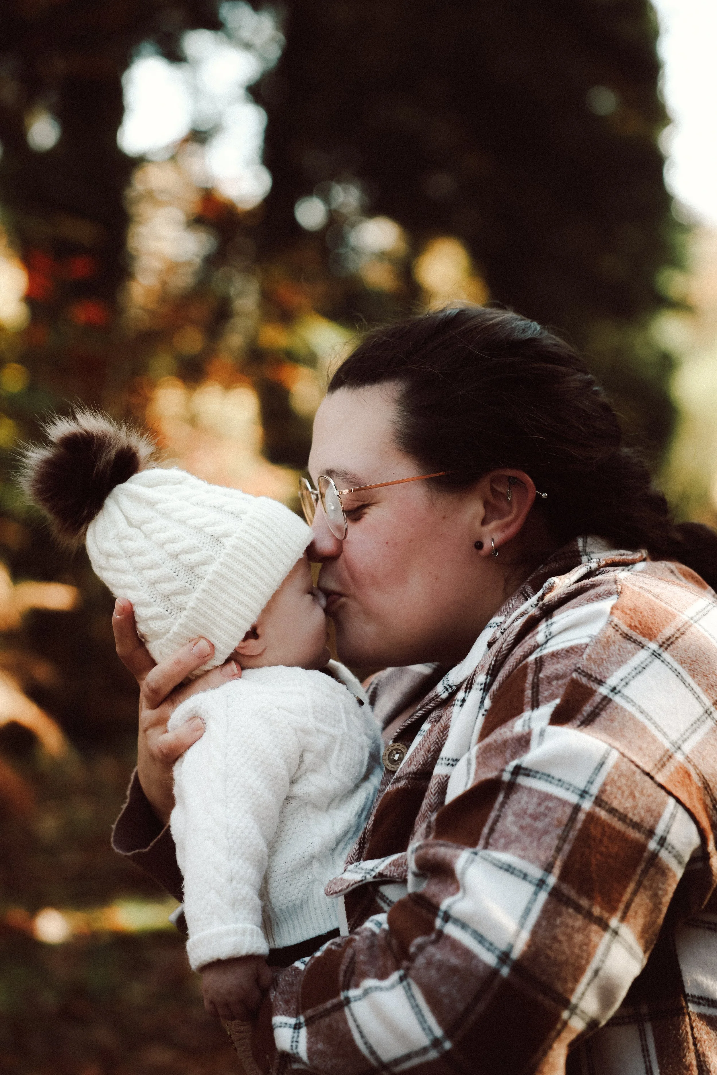 Une femme embrasse tendrement un jeune enfant dans un parc en automne, tous deux portant des vêtements chauds, avec un fond flou de feuilles d'arbres aux couleurs automnales.