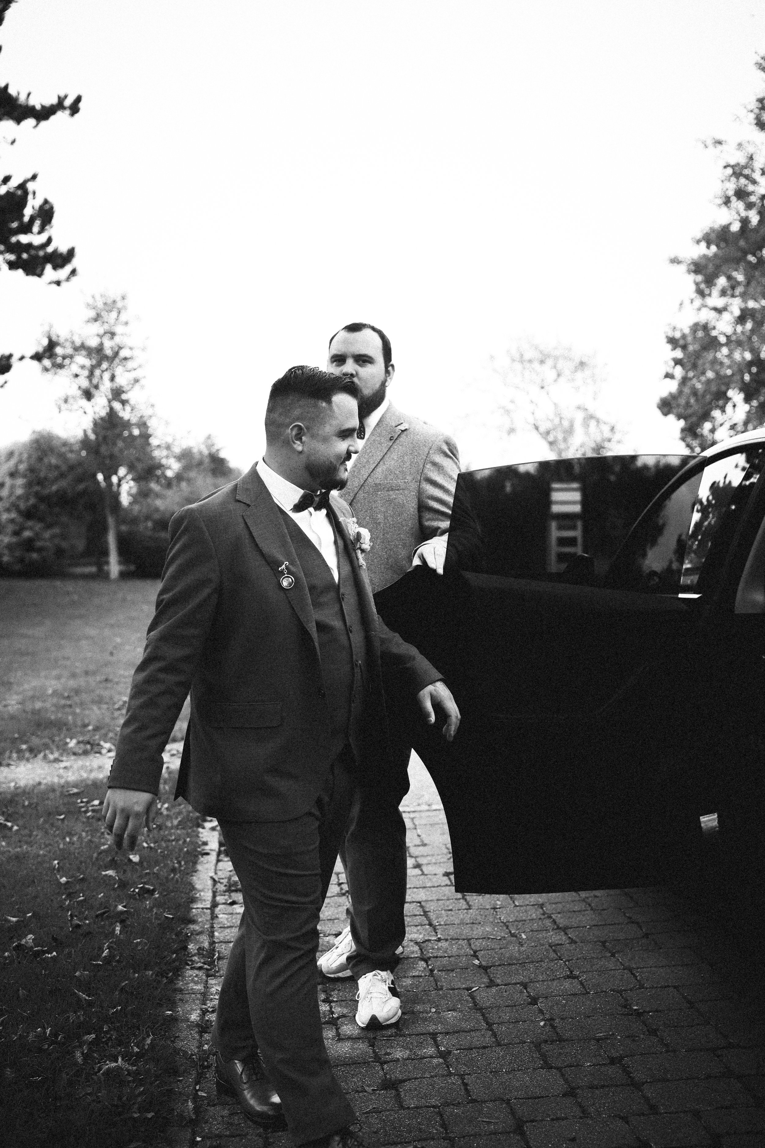 Deux hommes habillés en costumes de mariage devant une voiture noire.