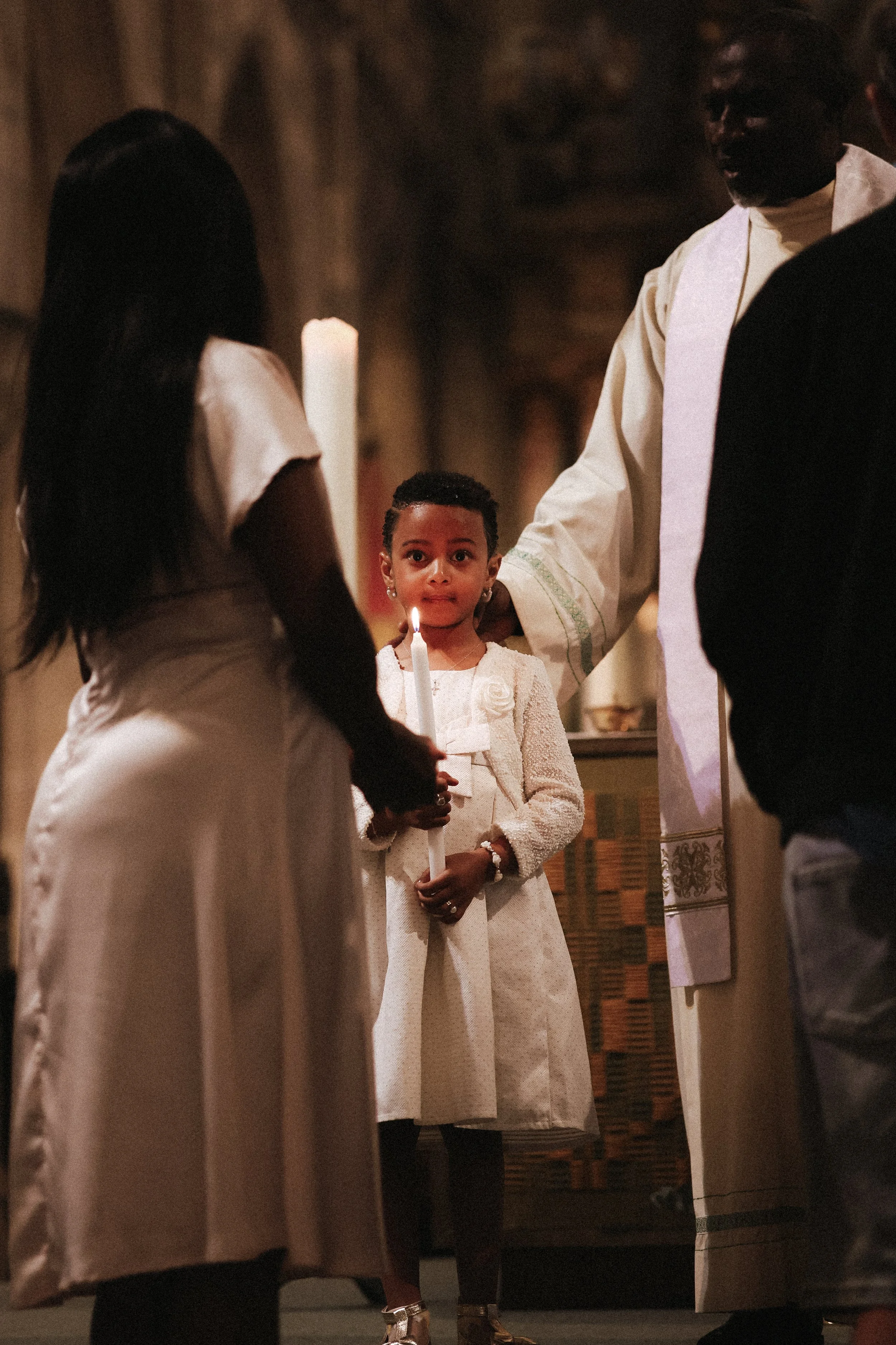 Une jeune fille lors d'une cérémonie religieuse, tenant une bougie allumée, entourée de personnes dont un homme en robe cérémonielle.