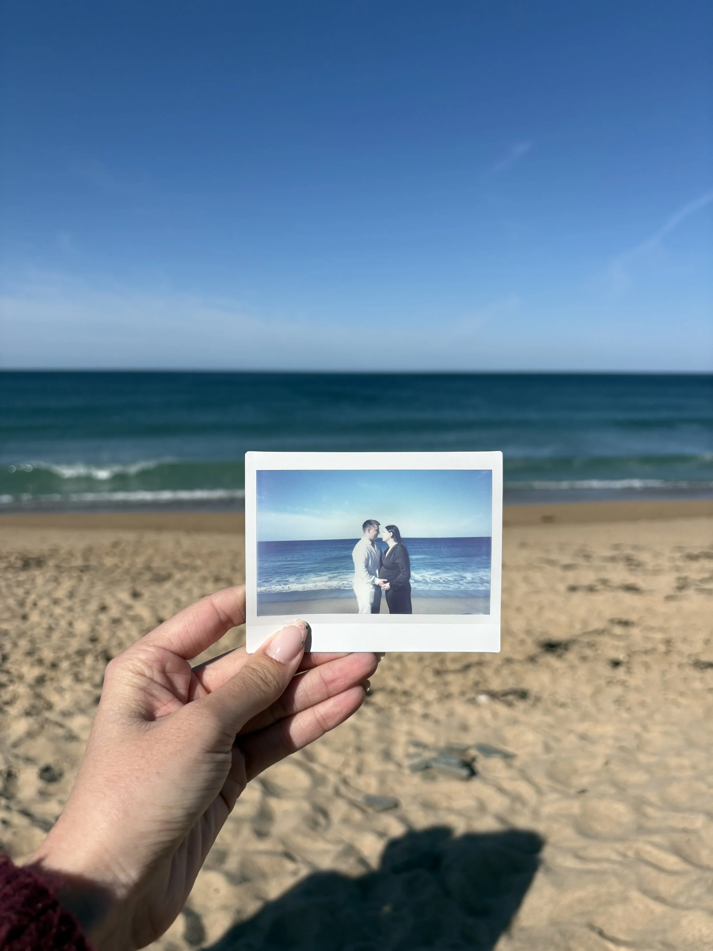 Une personne tient une photo instantanée d'un couple devant la mer, avec un ciel bleu et des plages de sable en arrière-plan.