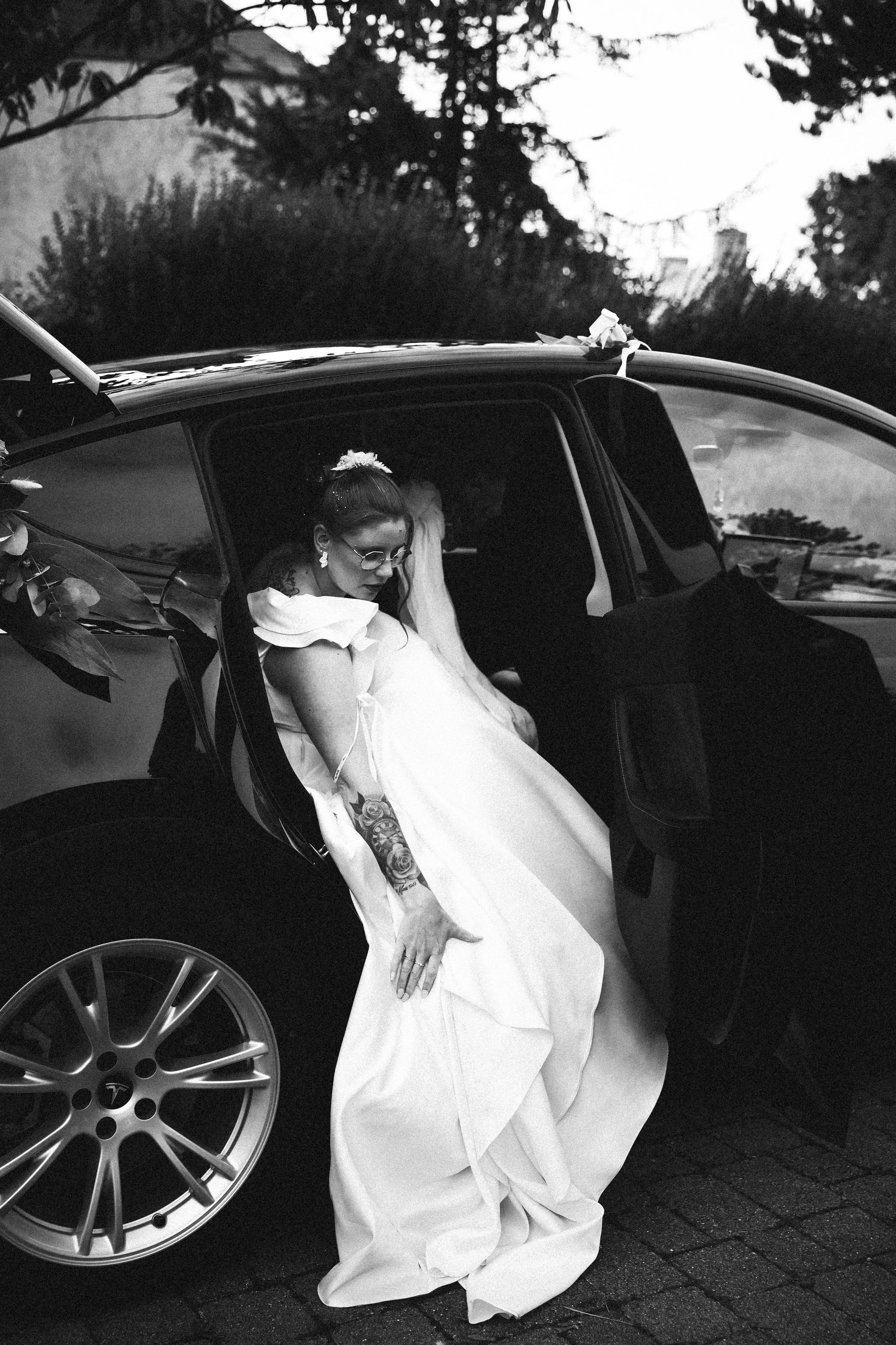 Une femme en robe de mariage sort d'une voiture noire, portant des lunettes et des boucles d'oreilles. Elle est assise à côté de la portière ouverte dans une ambiance urbaine.