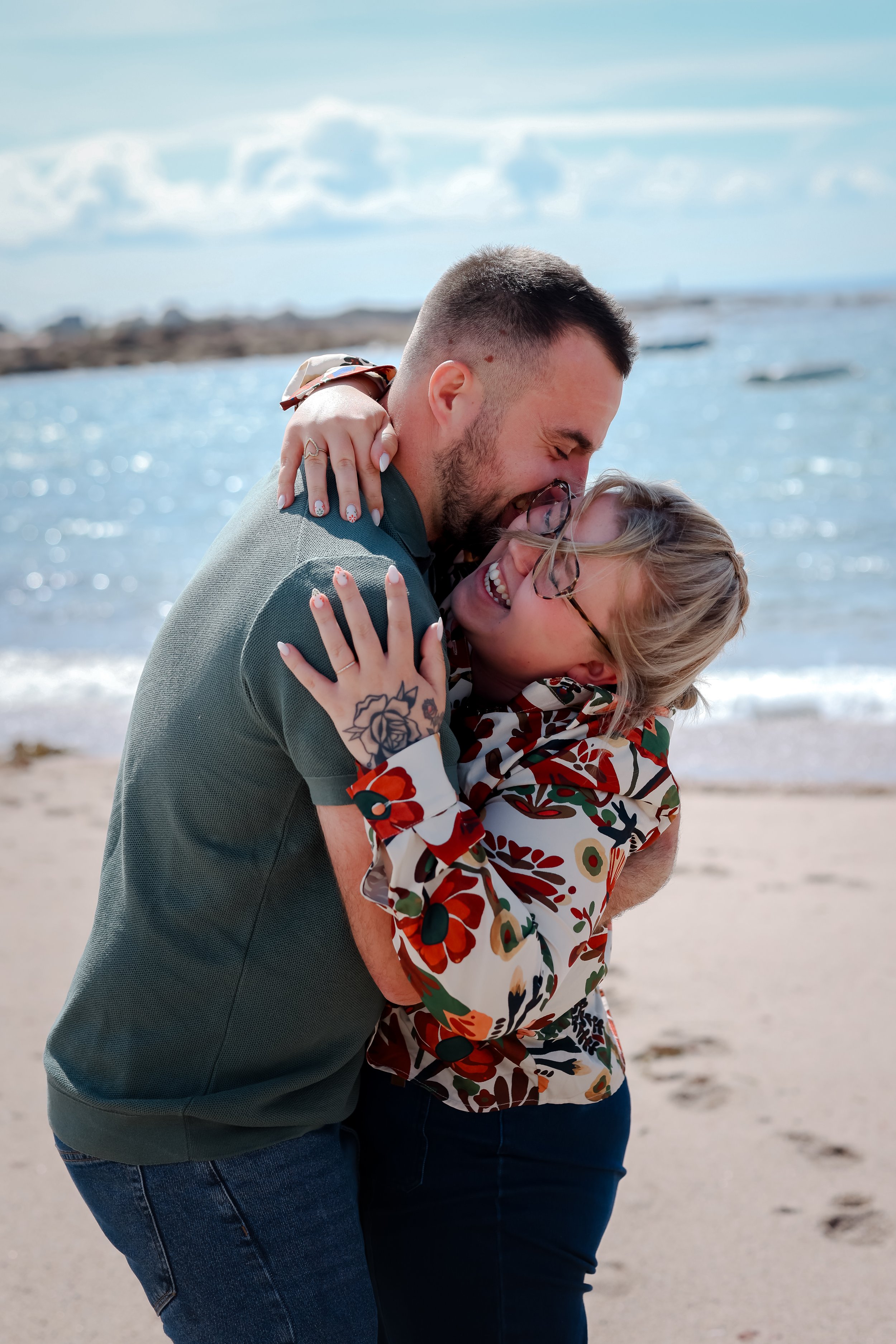 Un couple s'embrasse et rit sur la plage avec une mer en arrière-plan, vêtus de vêtements colorés. Il fait beau et ensoleillé.