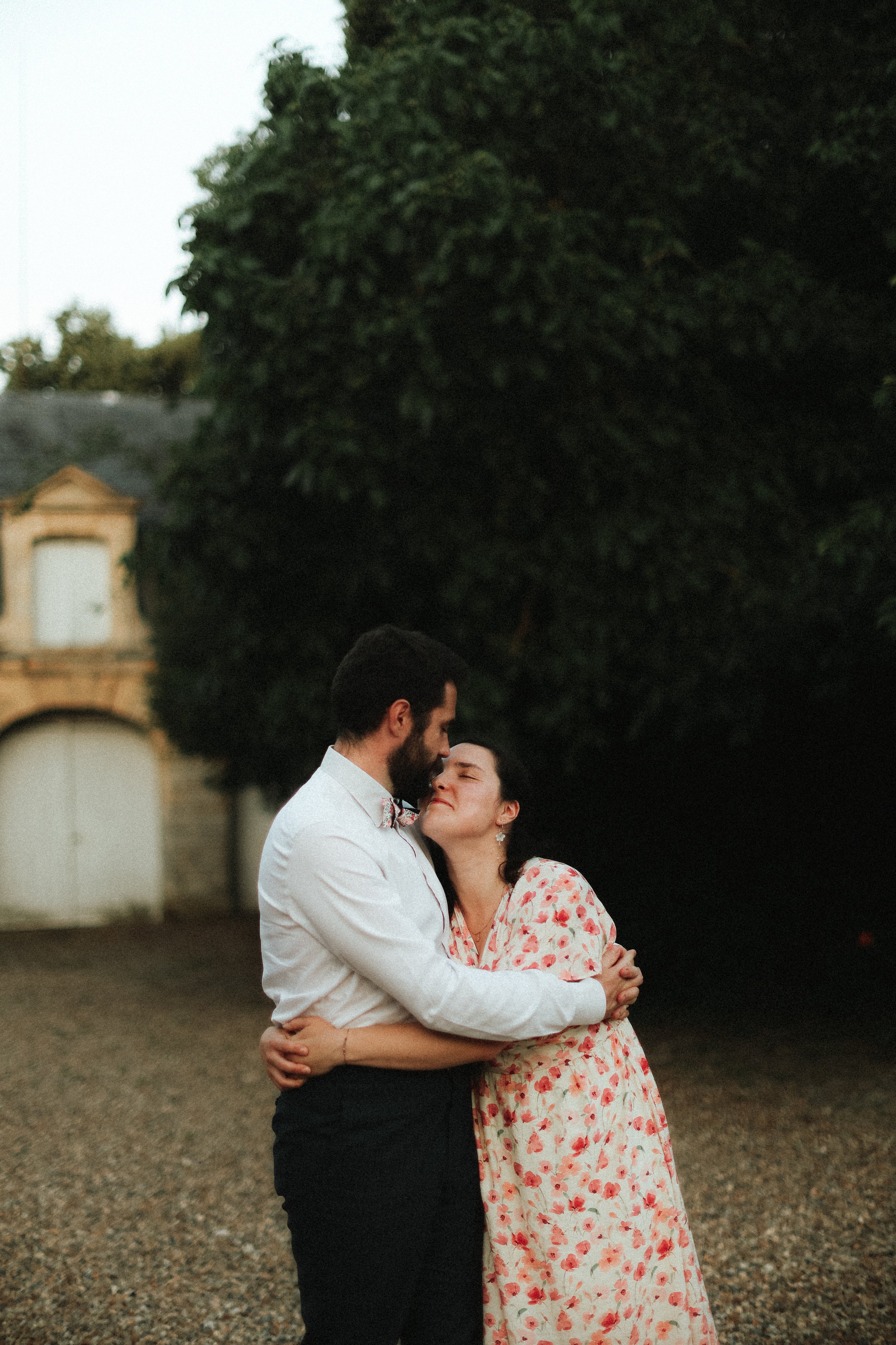 Un couple s'embrassant tendrement dehors, à l'approche de la nuit, avec un grand arbre et une vieille maison en arrière-plan.