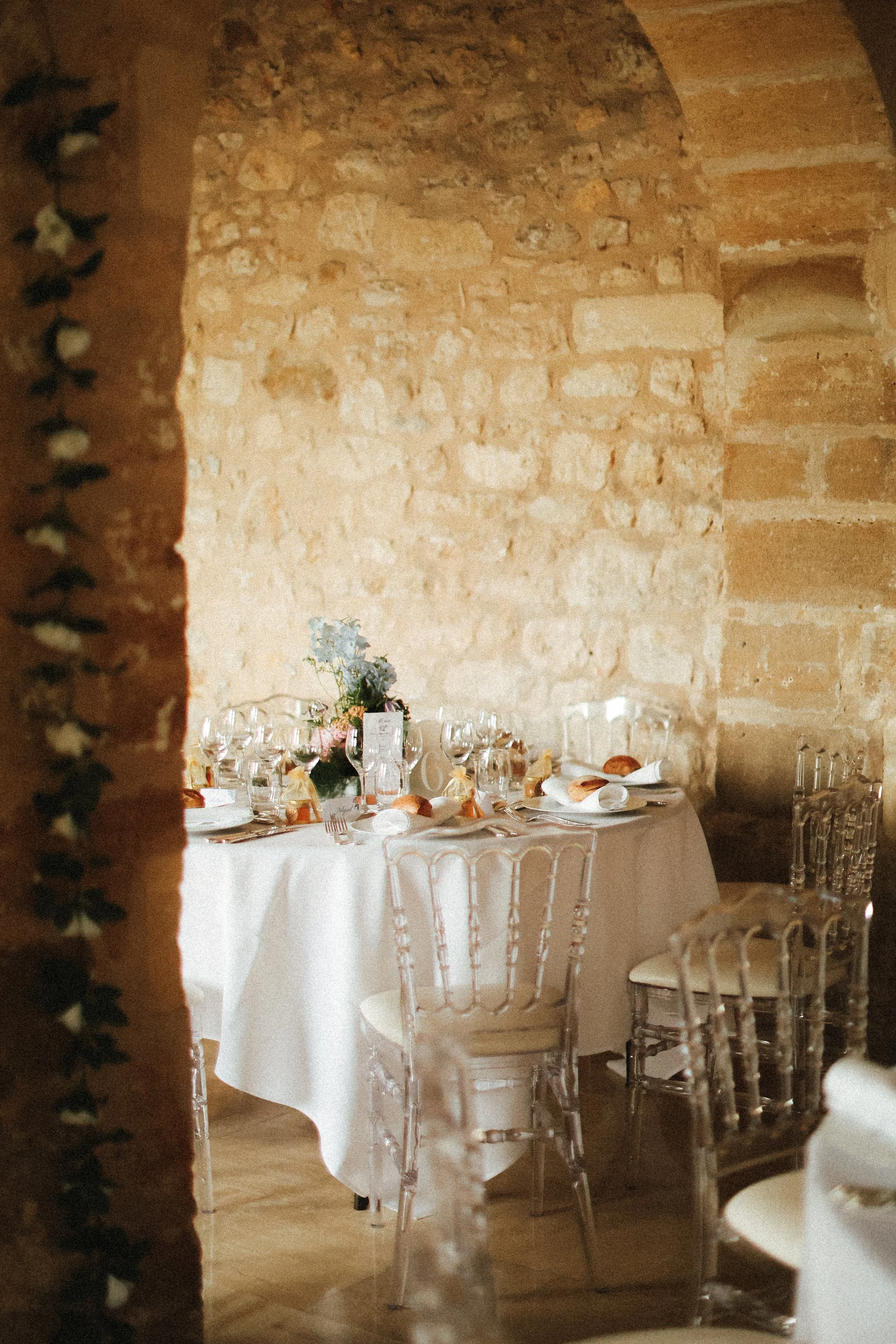 Salle de réception élégante avec un mur en pierre et une table ronde dressée pour un repas, ornée d'un centre de table floral et de verres à vin.