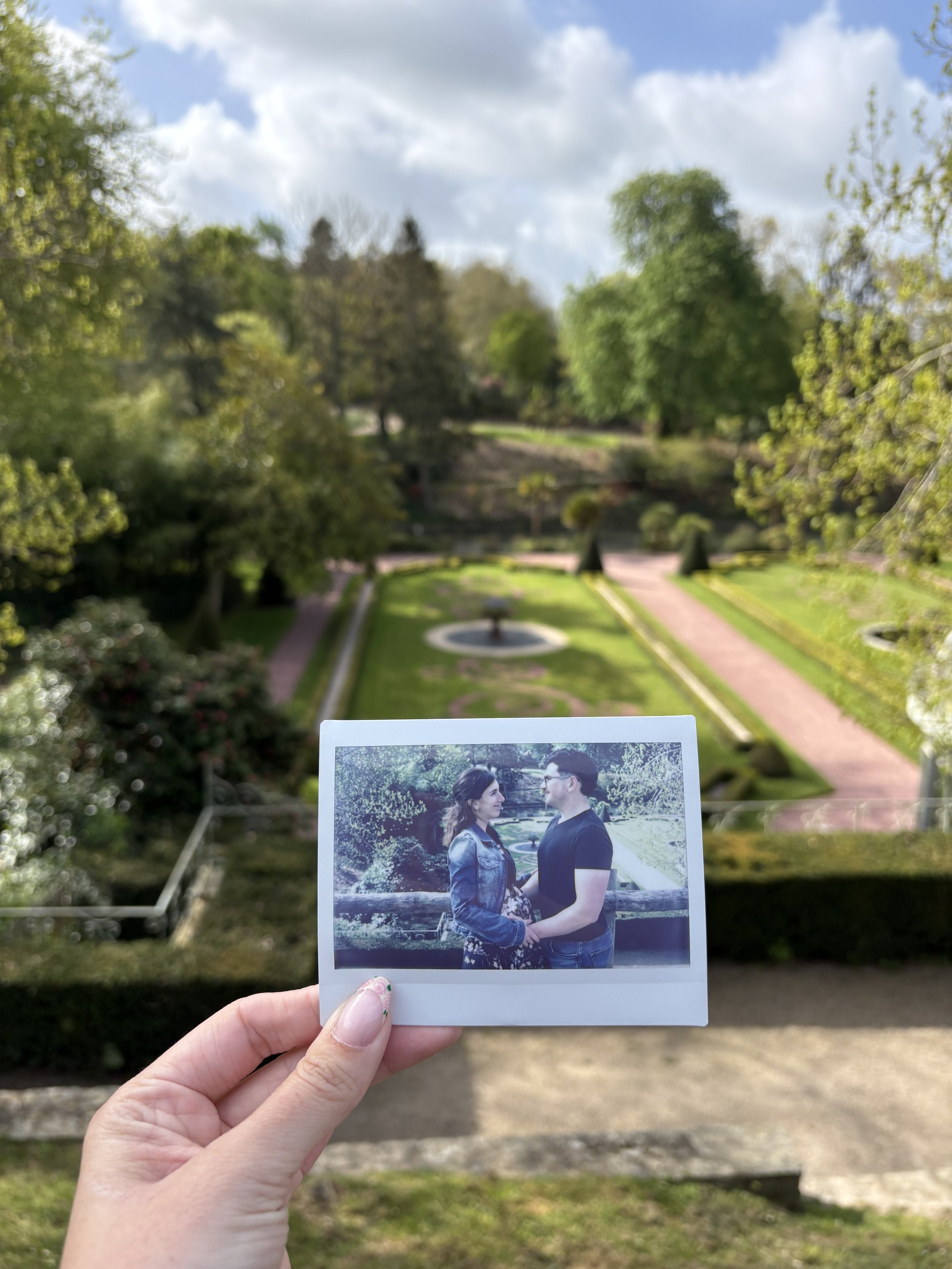 Une personne tient une photo polaroid d'un couple regardant dans les yeux, avec un parc verdoyant et un ciel partiellement nuageux à l'arrière-plan.