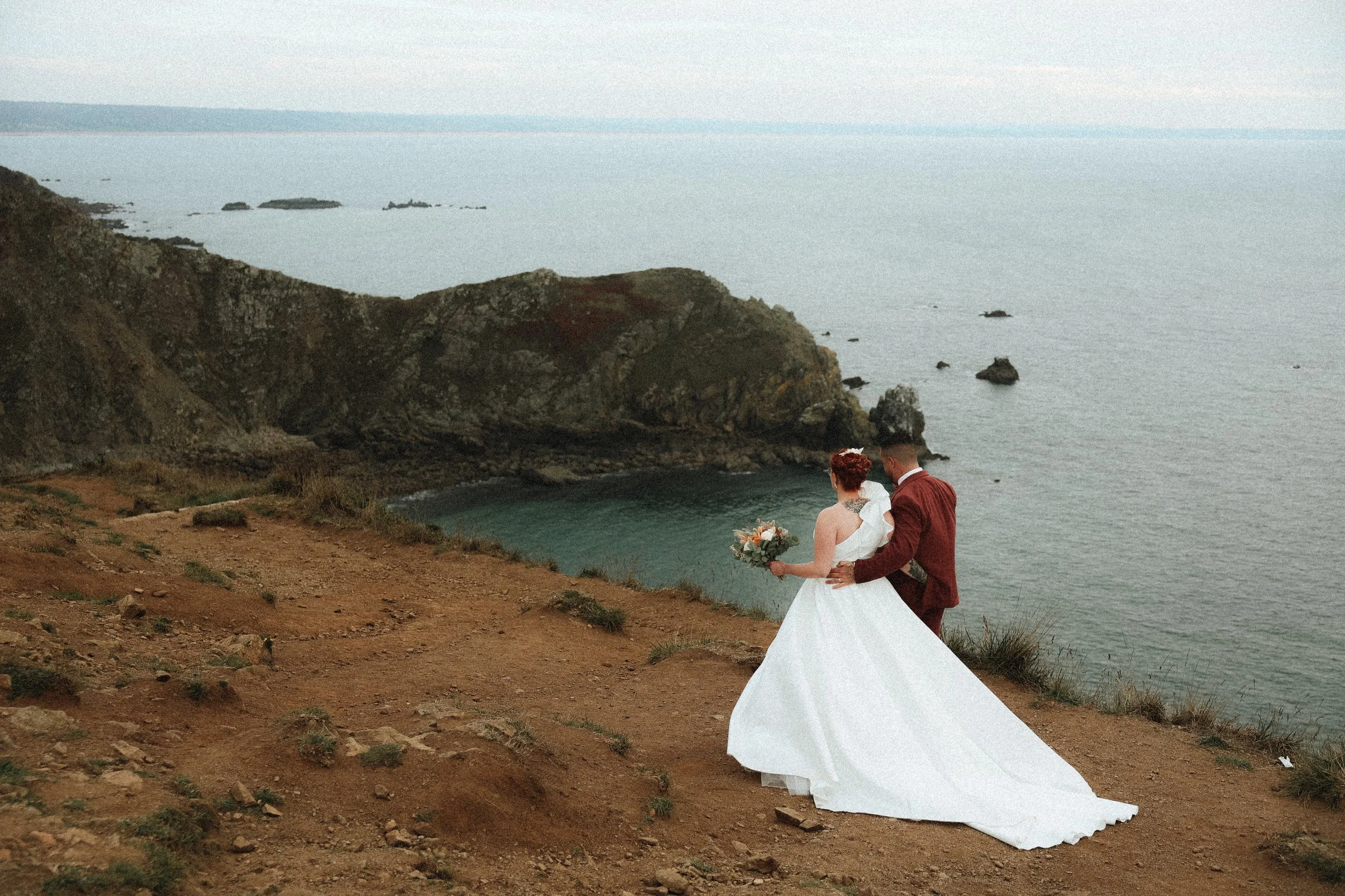 Un couple de mariés face à la mer, avec un décor rocheux et une côte, dans une robe de mariage blanche et un costume rouge.