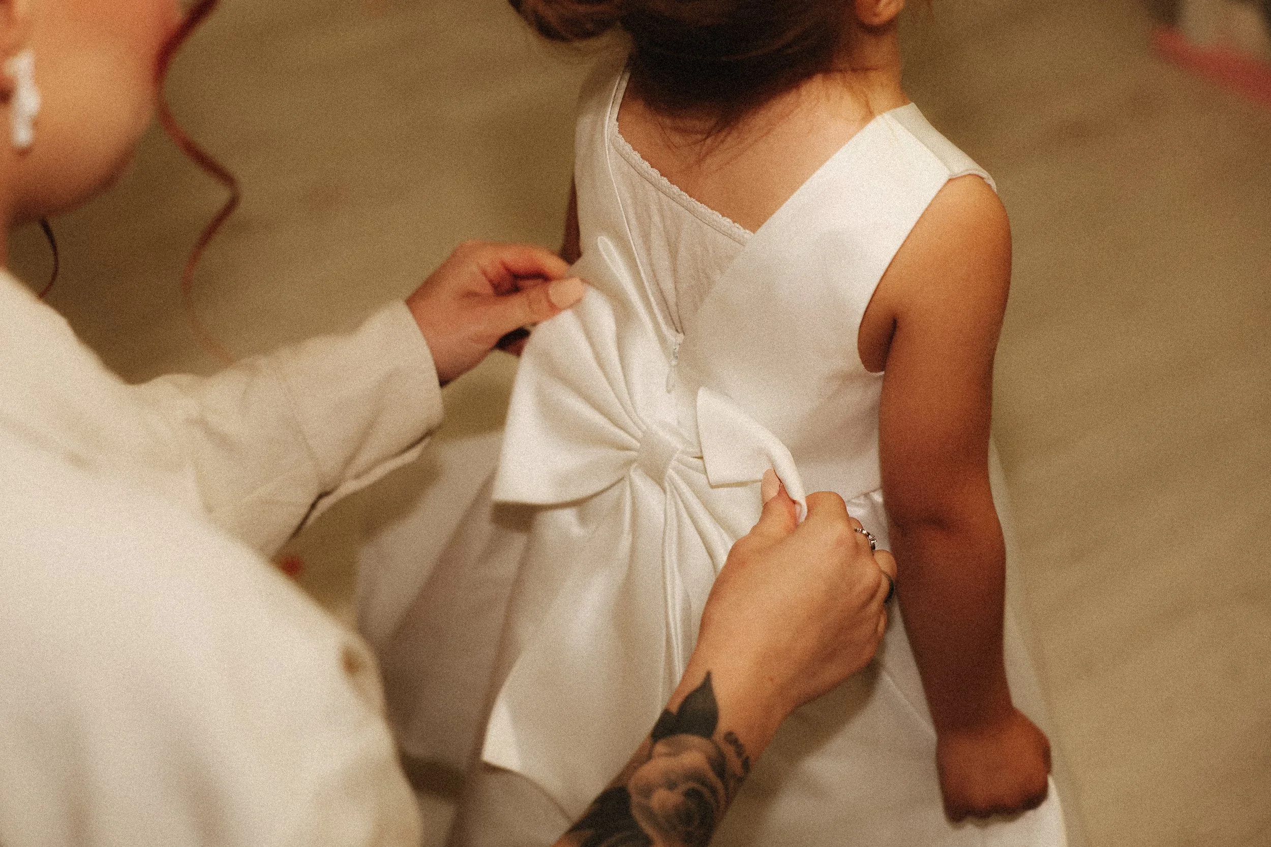 Une jeune fille en robe blanche est aidée à ajuster sa robe. La main de la personne qui l’aide montre un tatouage sur le bras.
