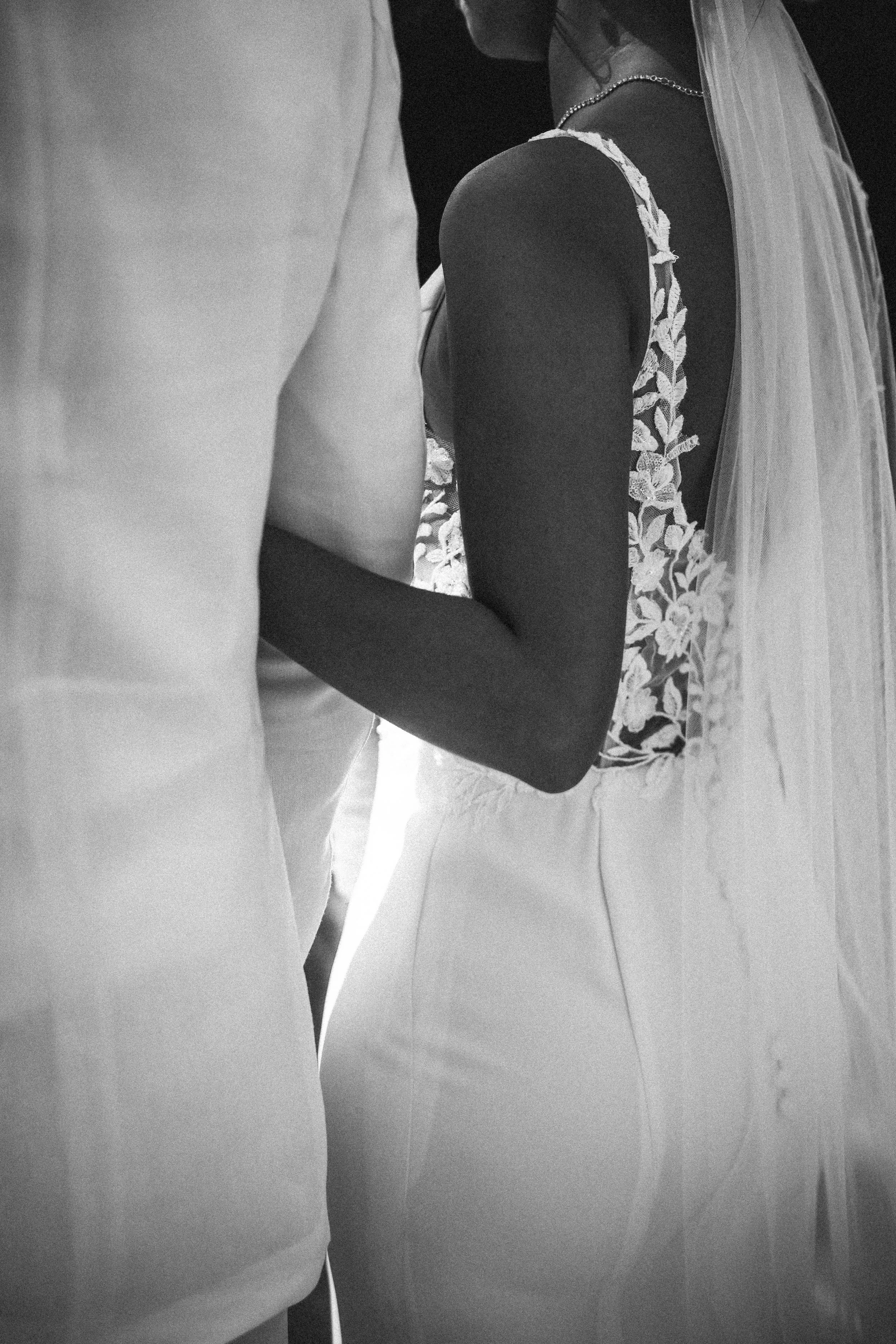 Une femme en robe blanche de mariage et un homme en costume lors d'une cérémonie nuptiale, vue de dos en noir et blanc.