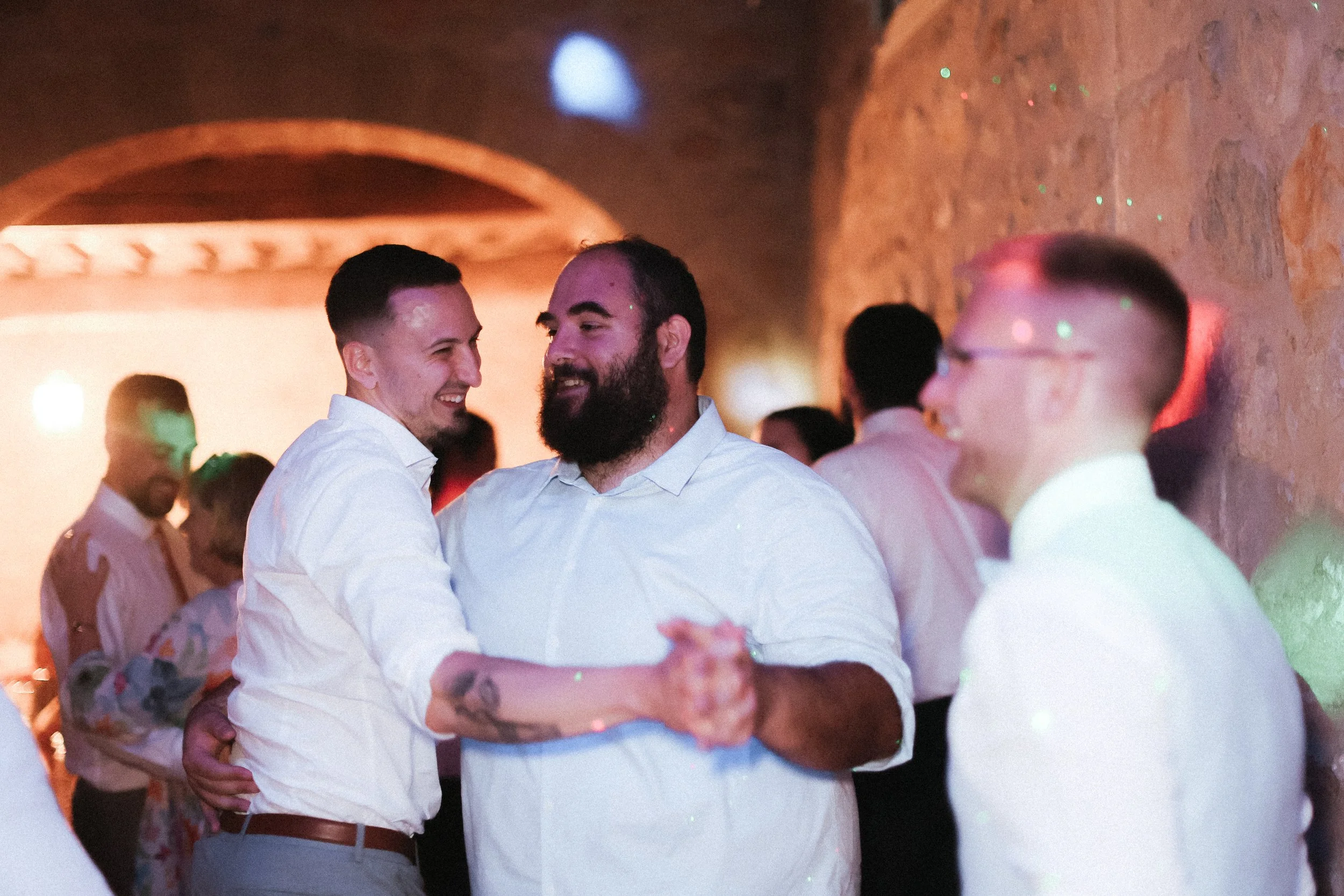 Deux hommes dansent et sourient lors d'une fête ou d'un mariage, entourés d'autres personnes, dans une ambiance chaleureuse et festive.