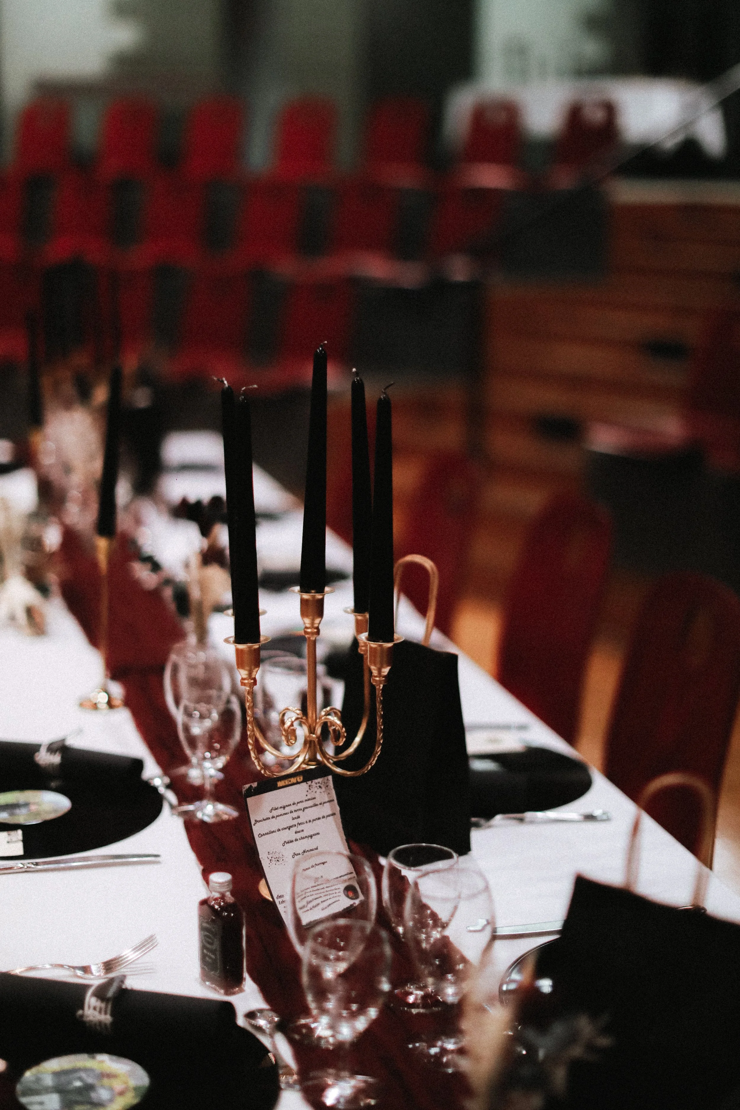 Table de fête décorée avec un chandelier noir et or, verres à vin, menus, et bouteilles, dans une salle avec des chaises rouges.