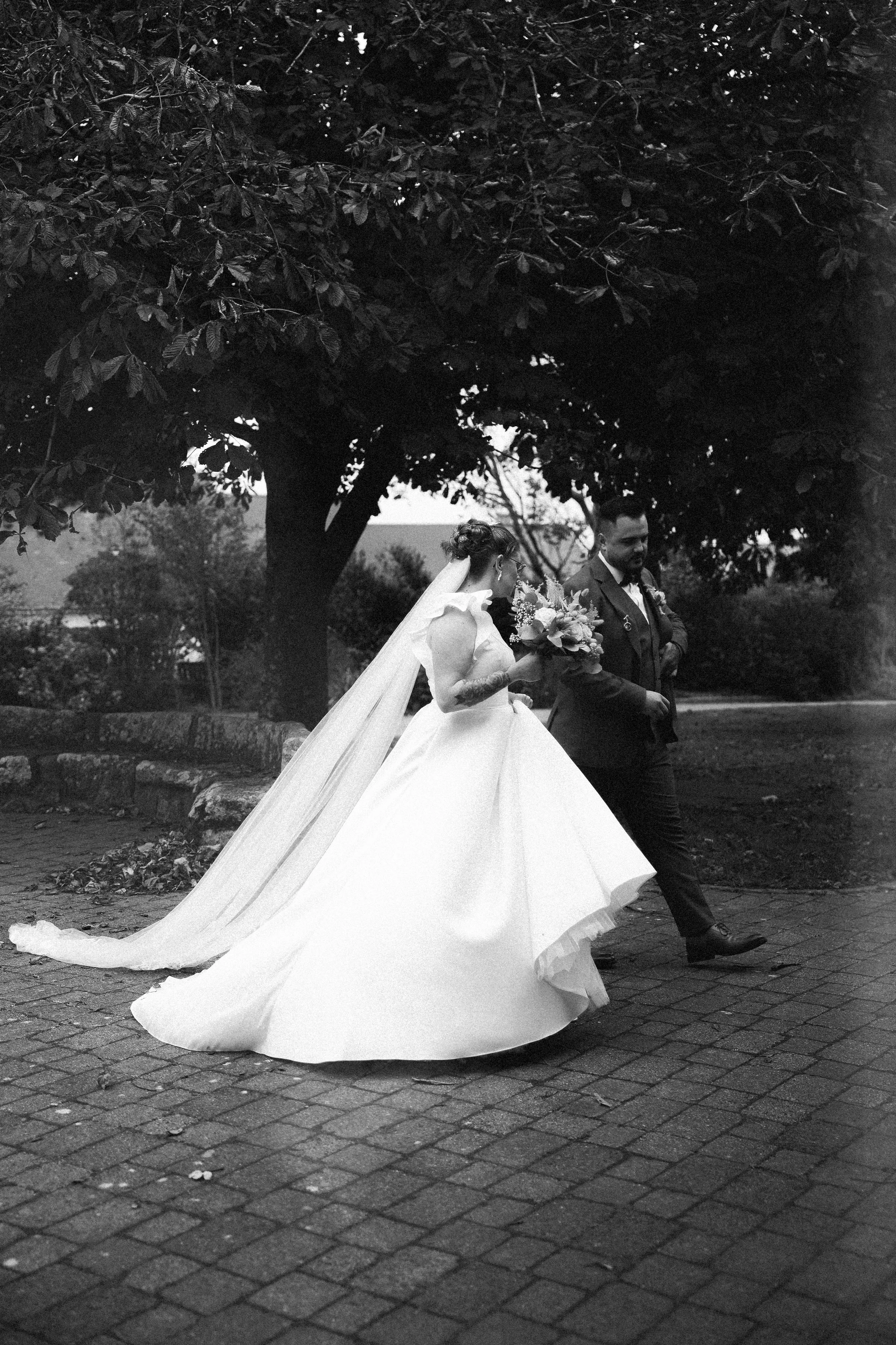 Une paire de mariés marche dans un parc, la mariée porte une robe de mariée blanche et tient un bouquet, le marié porte un costume sombre, tous deux sous un grand arbre en arrière-plan.