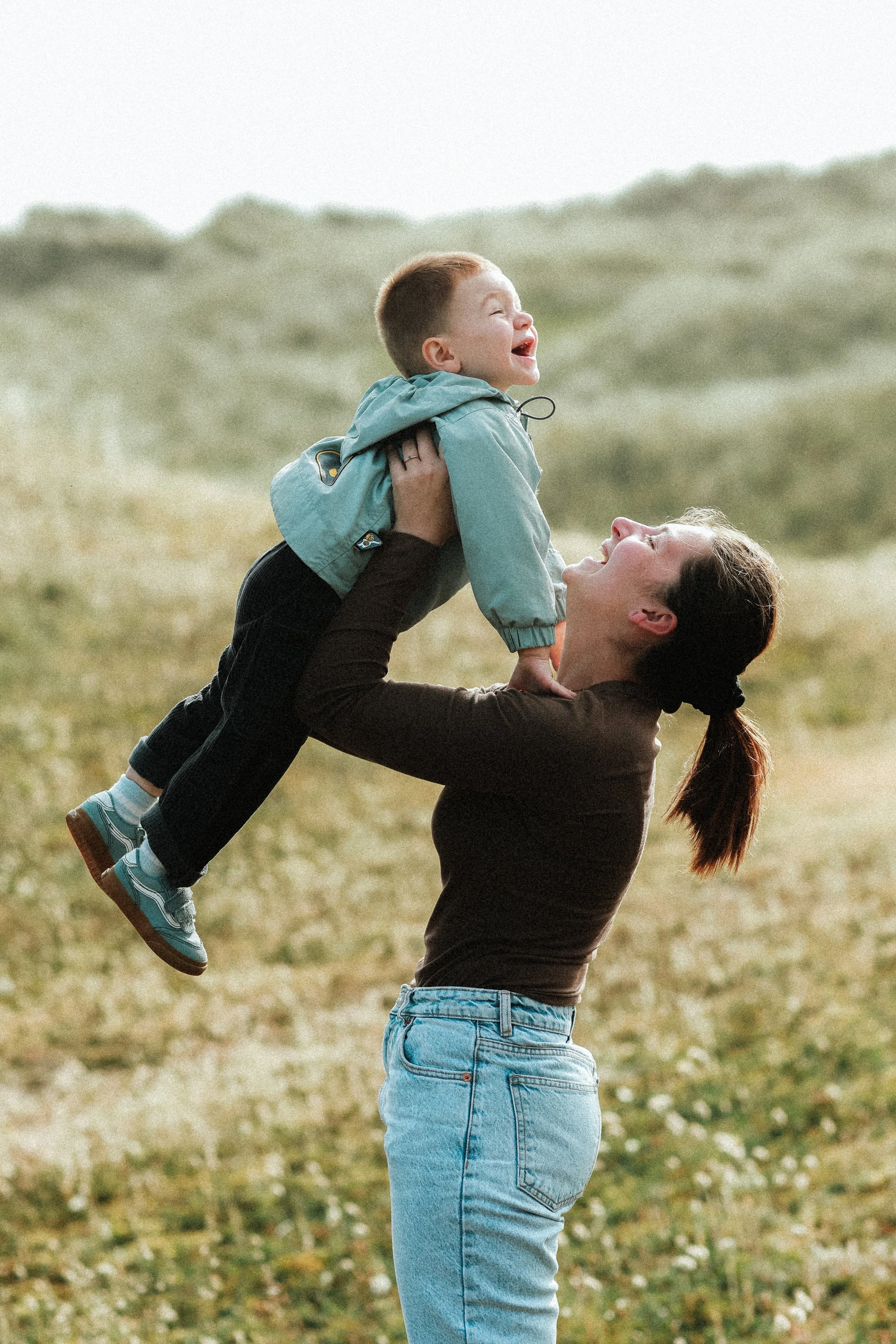 Une femme souriante soulève un enfant à l'extérieur, dans un champ avec des collines en arrière-plan.