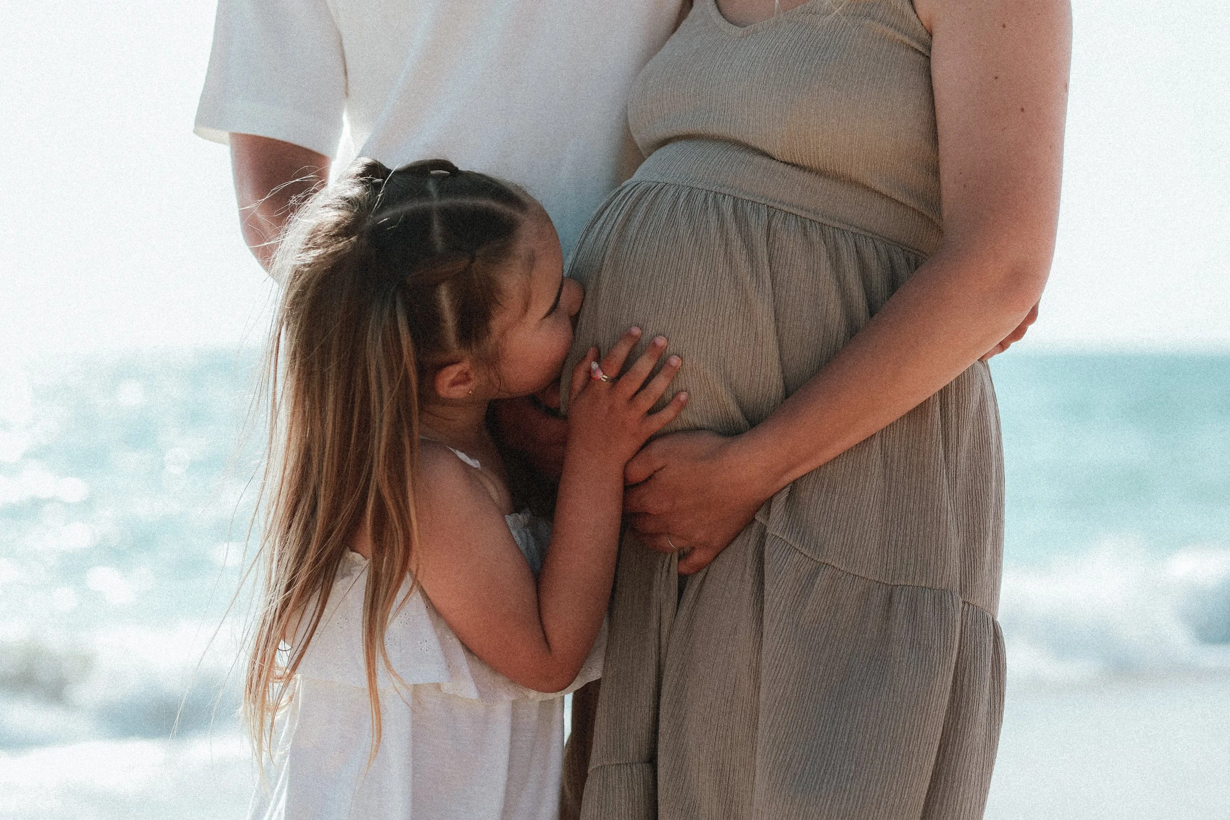 Une jeune fille embrasse le ventre d'une femme enceinte sur la plage, dans un cadre ensoleillé