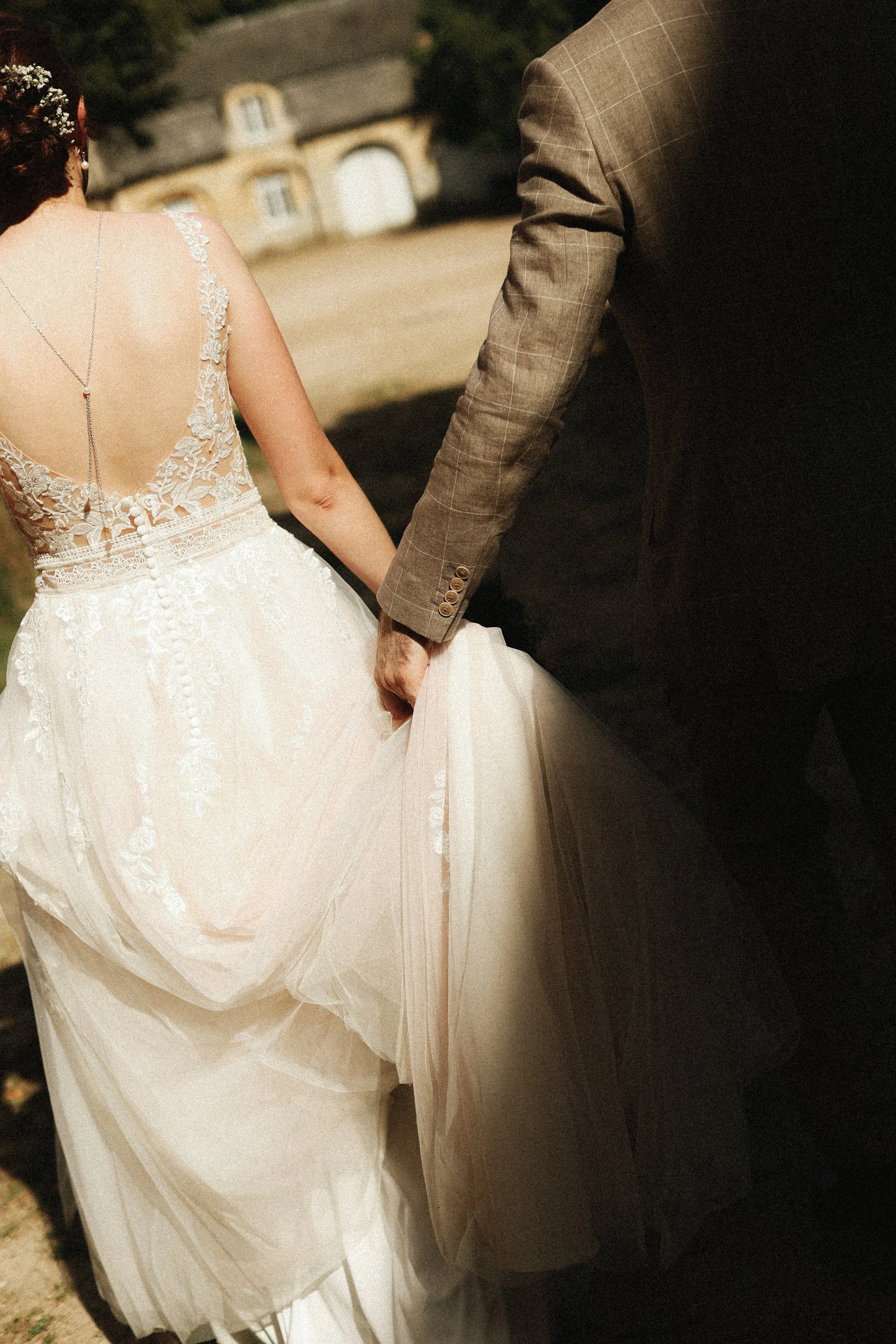 Un couple de mariés en costume et robe de mariage, main dans la main, à l'extérieur avec des bâtiments en pierre en arrière-plan.