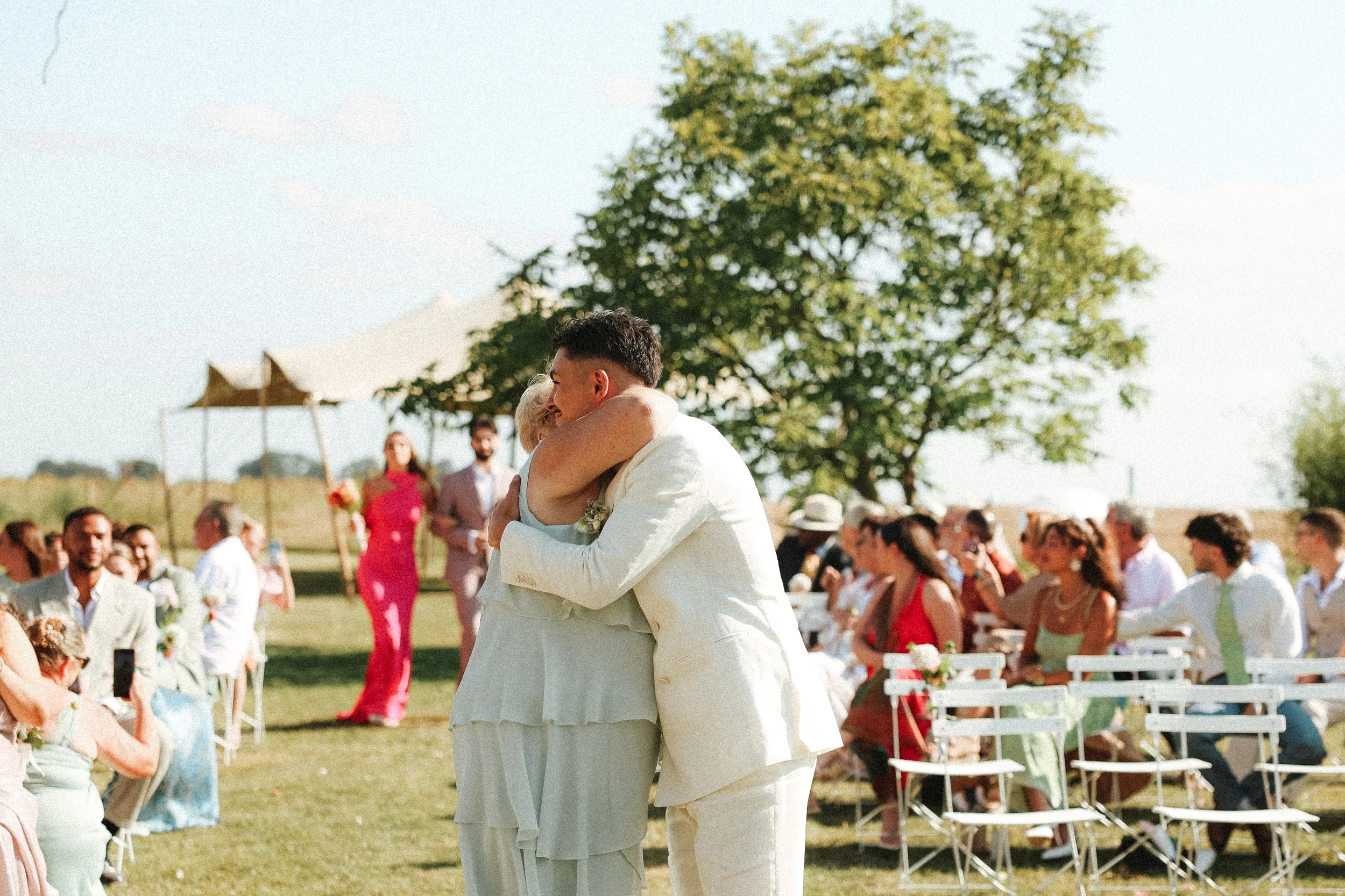Deux personnes en tenue de mariage s'étreignent lors d'une cérémonie en plein air, avec des invités assis et des arbres en arrière-plan.