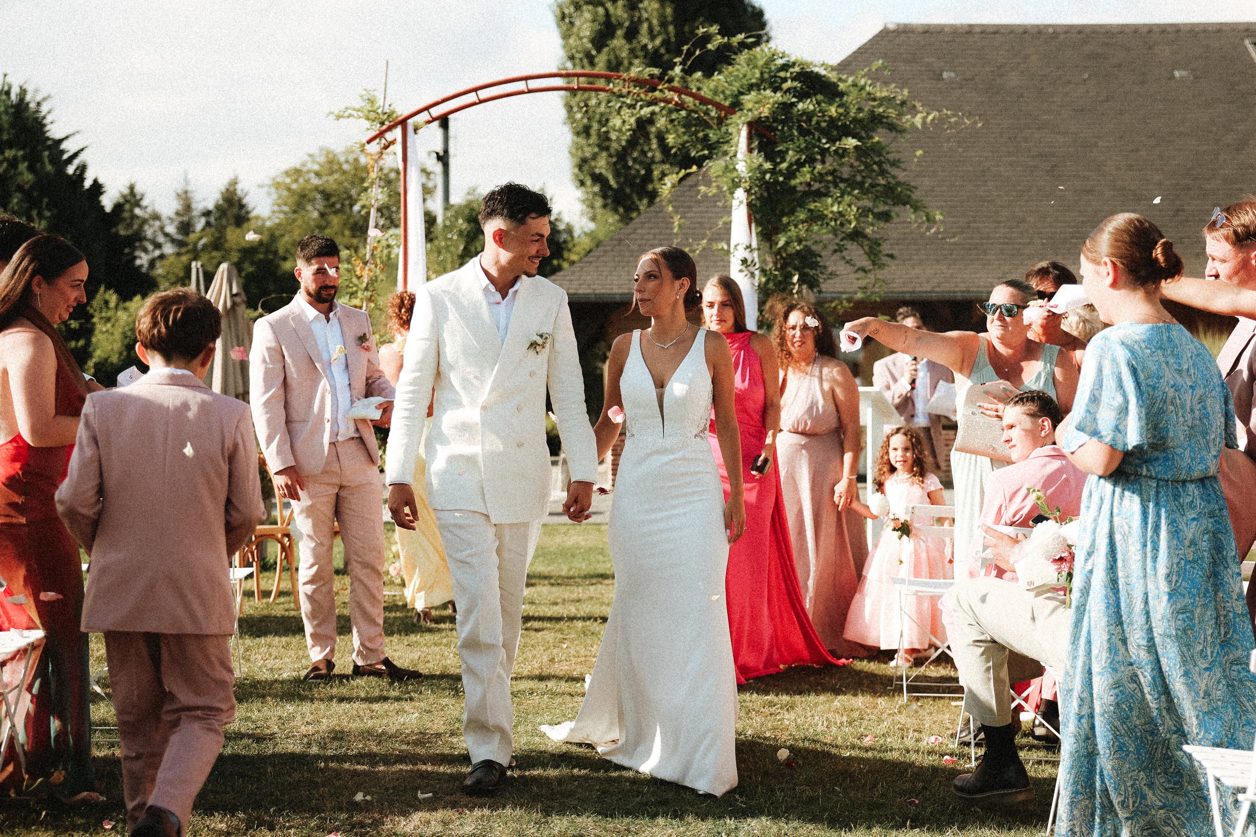 Un couple de mariés marche main dans la main au milieu d'une fête en plein air, entourés d'invités. La scène se déroule sur une pelouse avec des décorations florales et un arc d'arche en arrière-plan, par une journée ensoleillée.