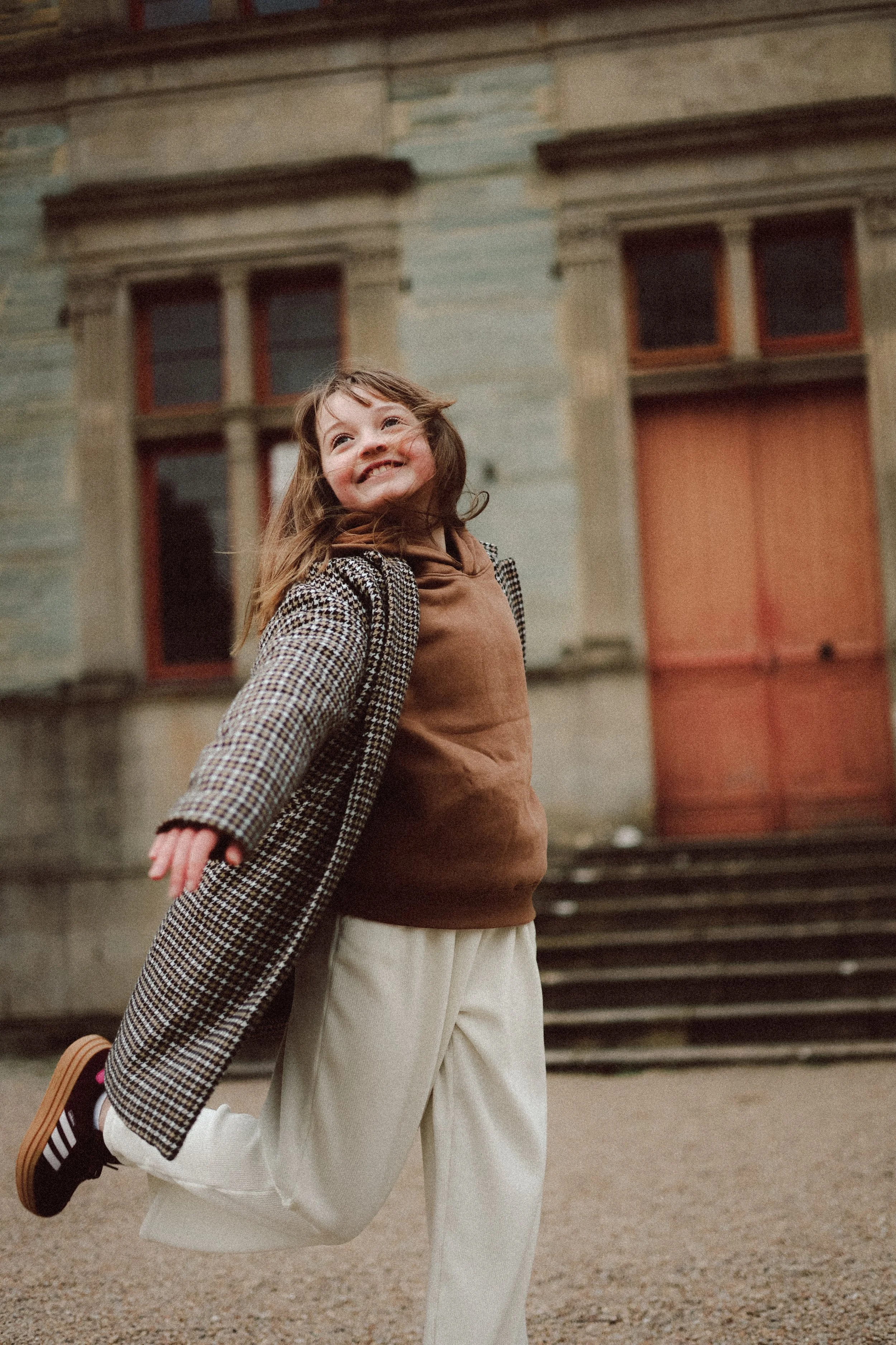 Une jeune fille souriante qui court devant un bâtiment ancien en pierre, portant un manteau à carreaux, un sweat marron, un pantalon beige et des chaussures noires.