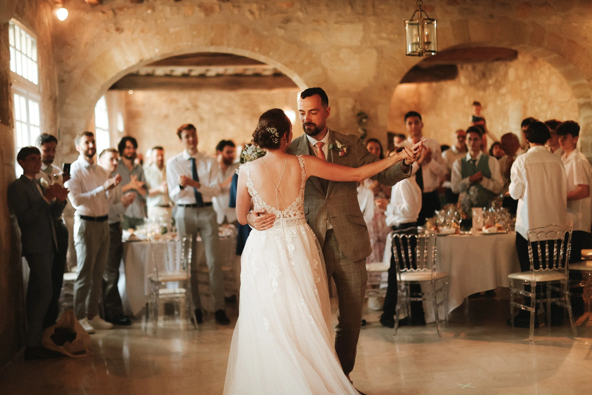 Un couple de mariés danse lors de leur réception de mariage dans une salle avec murs en pierre et grands fenêtres. Autour, des invités regardent et applaudissent, certains prennent des photos.