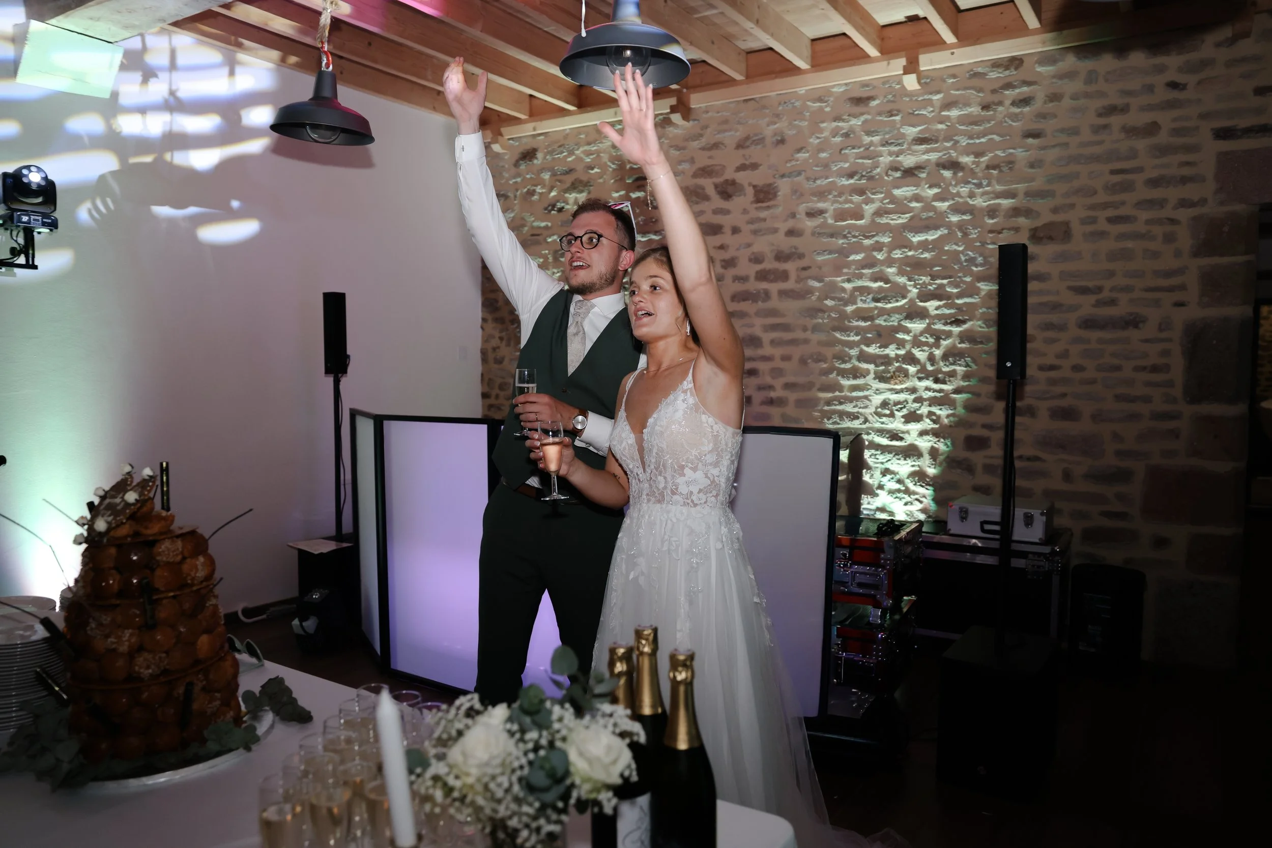 Un couple de mariés danse lors d'une réception dans une salle avec murs en pierre et plafond en bois, avec des décorations florales et une table de dessert en avant-plan.