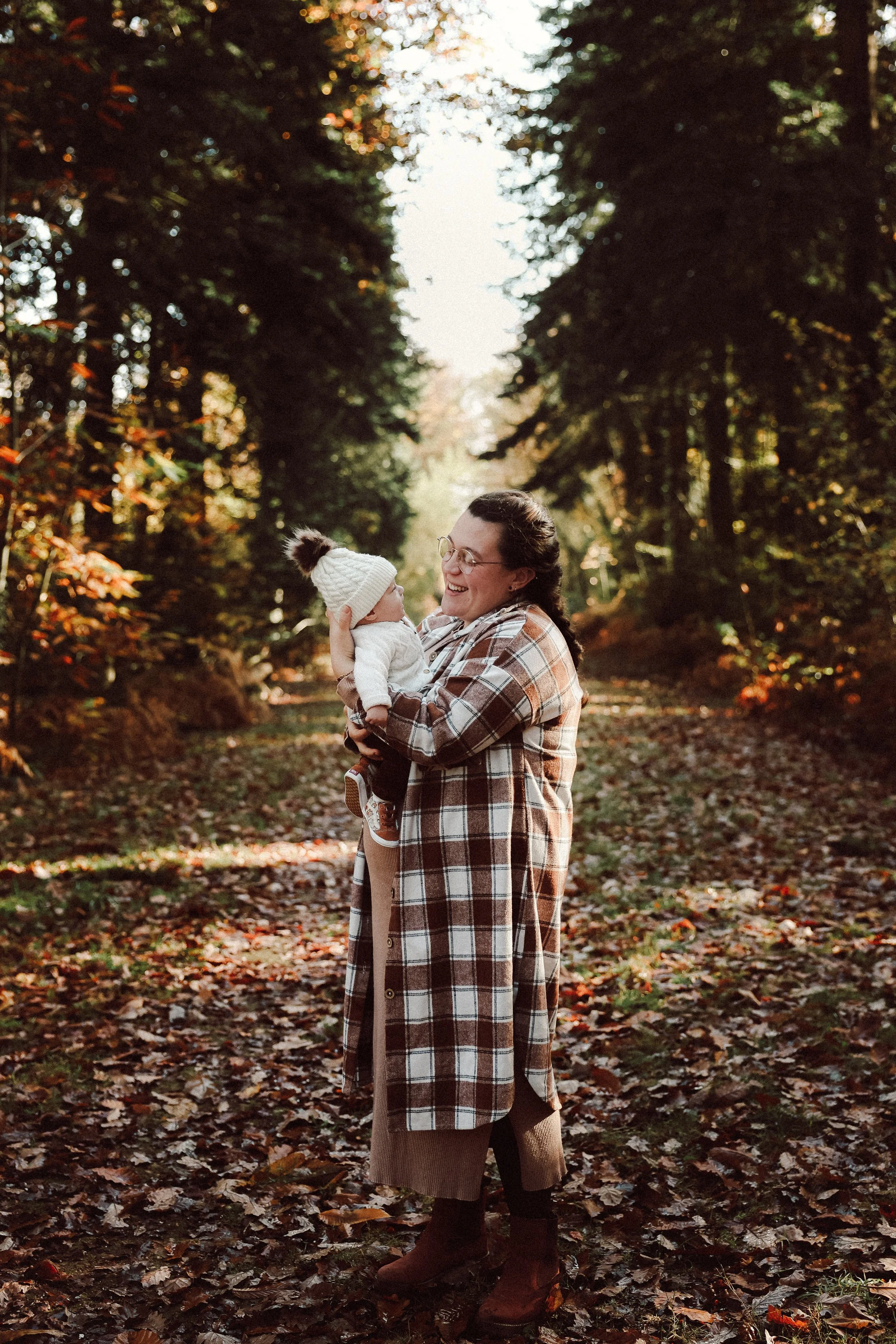 Une femme tenant un bébé dans une forêt en automne, tous deux souriants.