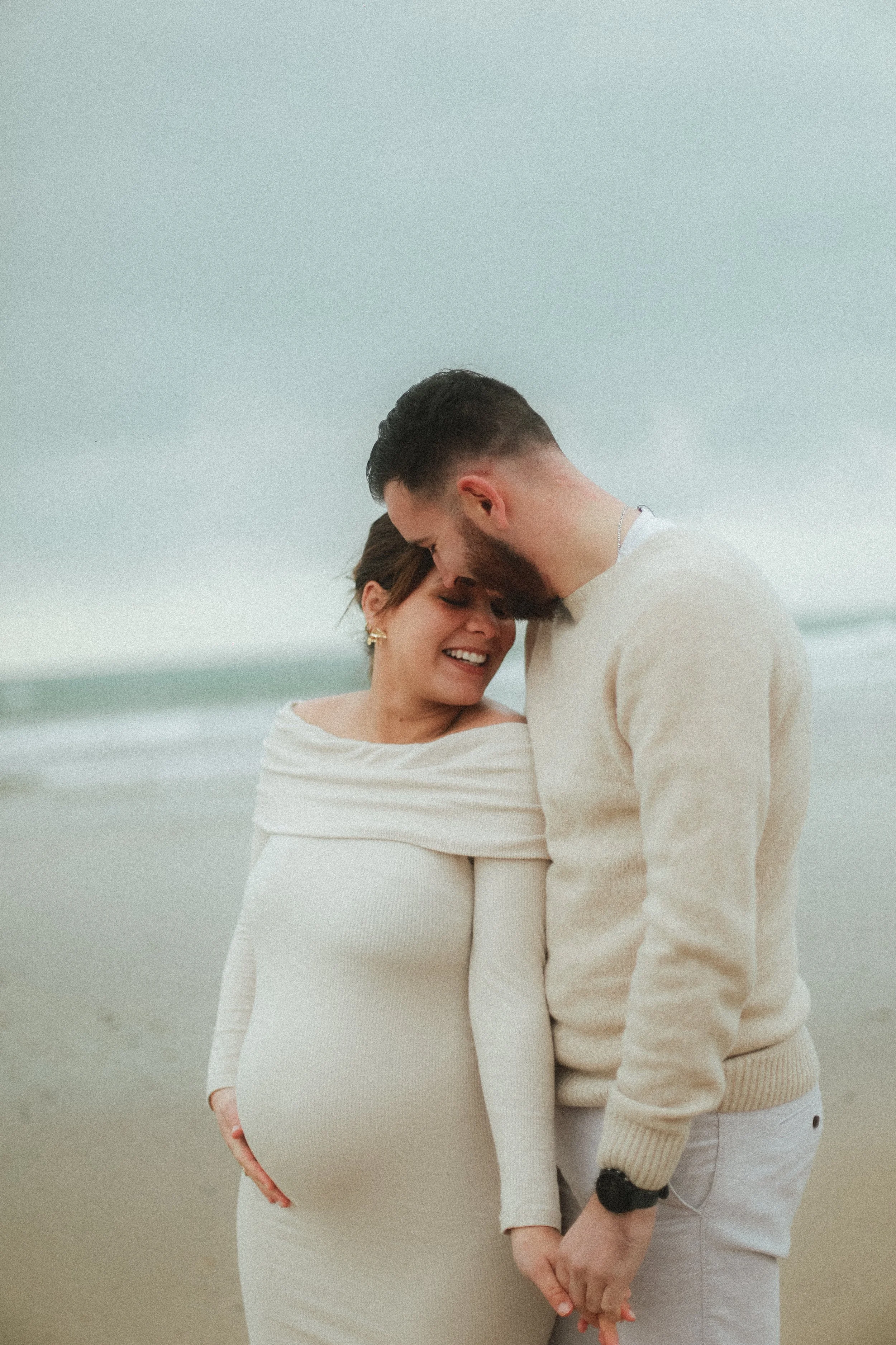 Un couple tenant la main sur la plage, un homme embrassant le front d'une femme enceinte, tous deux souriants et vêtus de blanc.