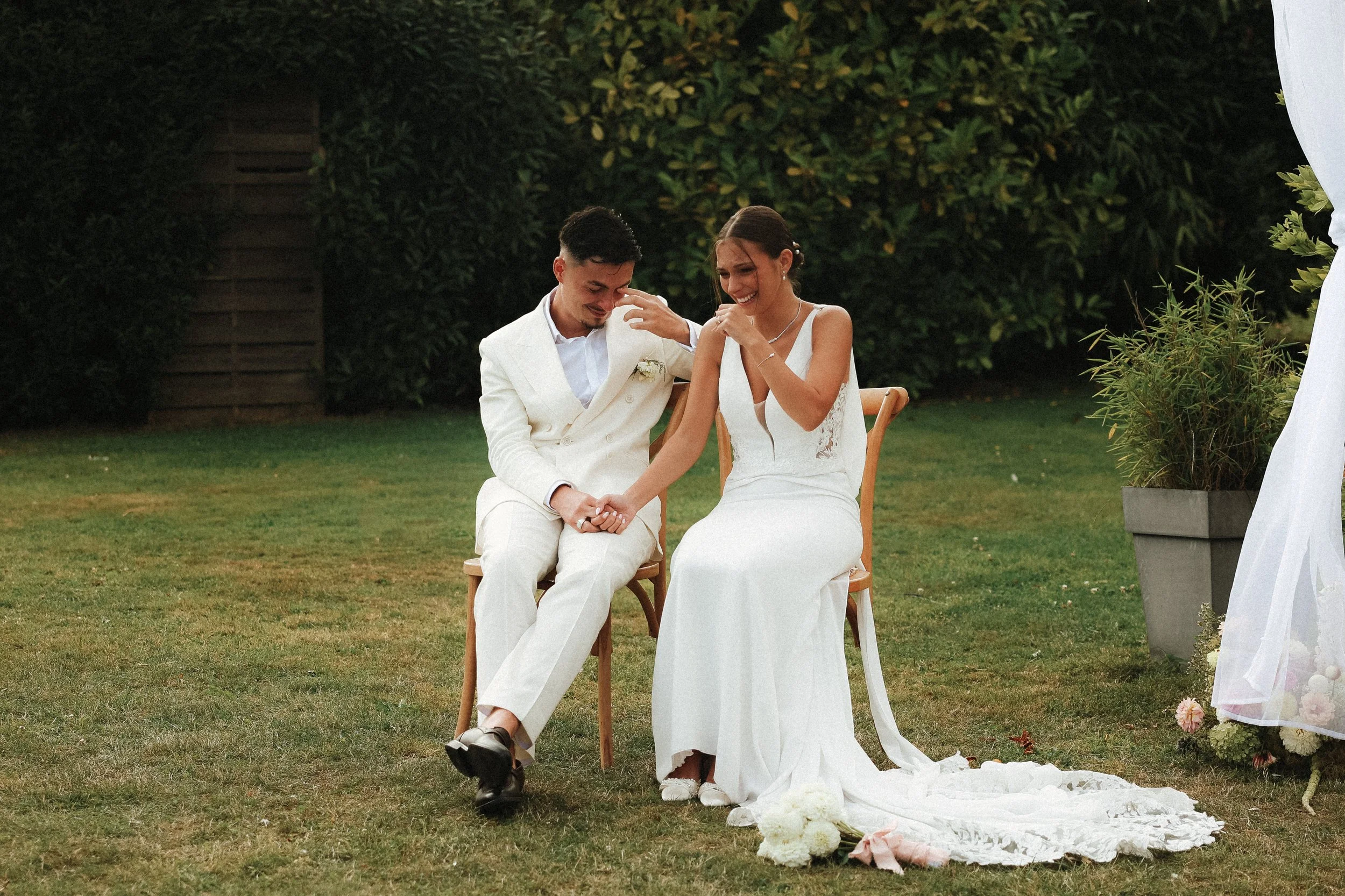 Un couple lors de leur mariage, assis dans un jardin, la femme en robe blanche, le mari en costume blanc, ils semblent émus, en train de s'embrasser ou de se consoler, entourés de verdure.