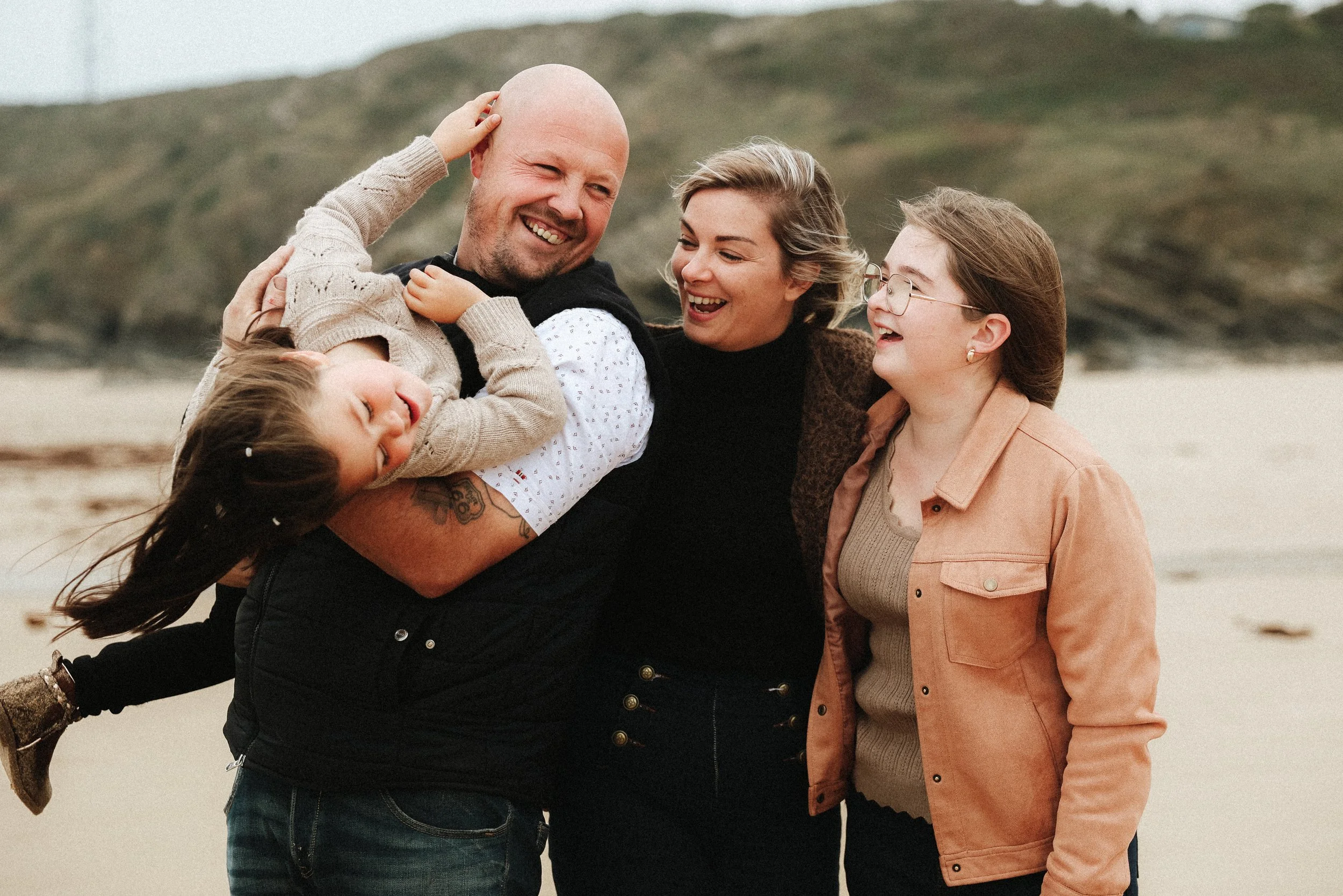 Un groupe de cinq personnes, dont une enfant, souriant et se tenant par les épaules, à la plage avec des collines en arrière-plan.