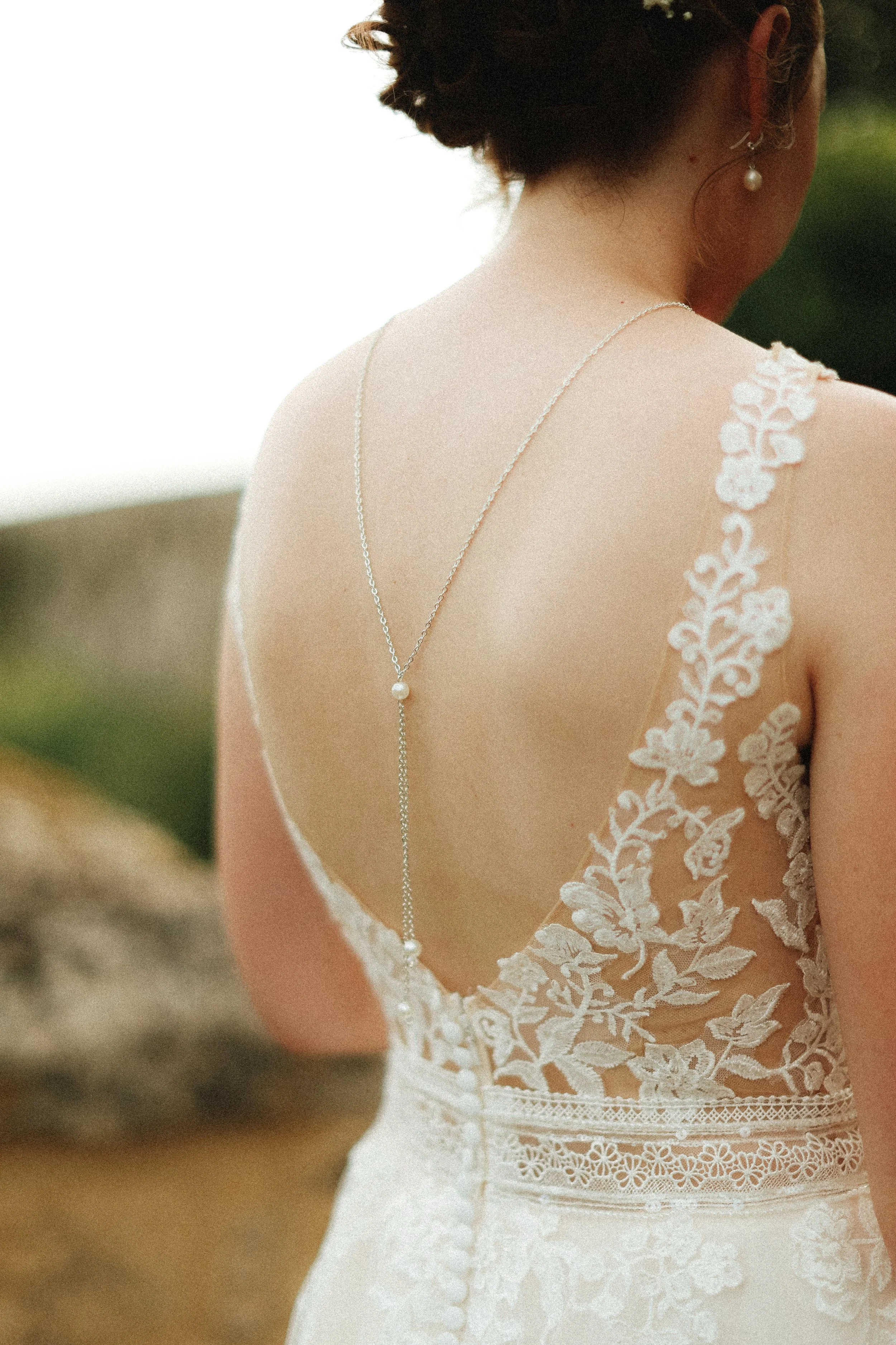 Une femme portant une robe de mariée en dentelle blanche, vue de dos, avec un collier fin et une boucle d'oreille en perle, en extérieur.